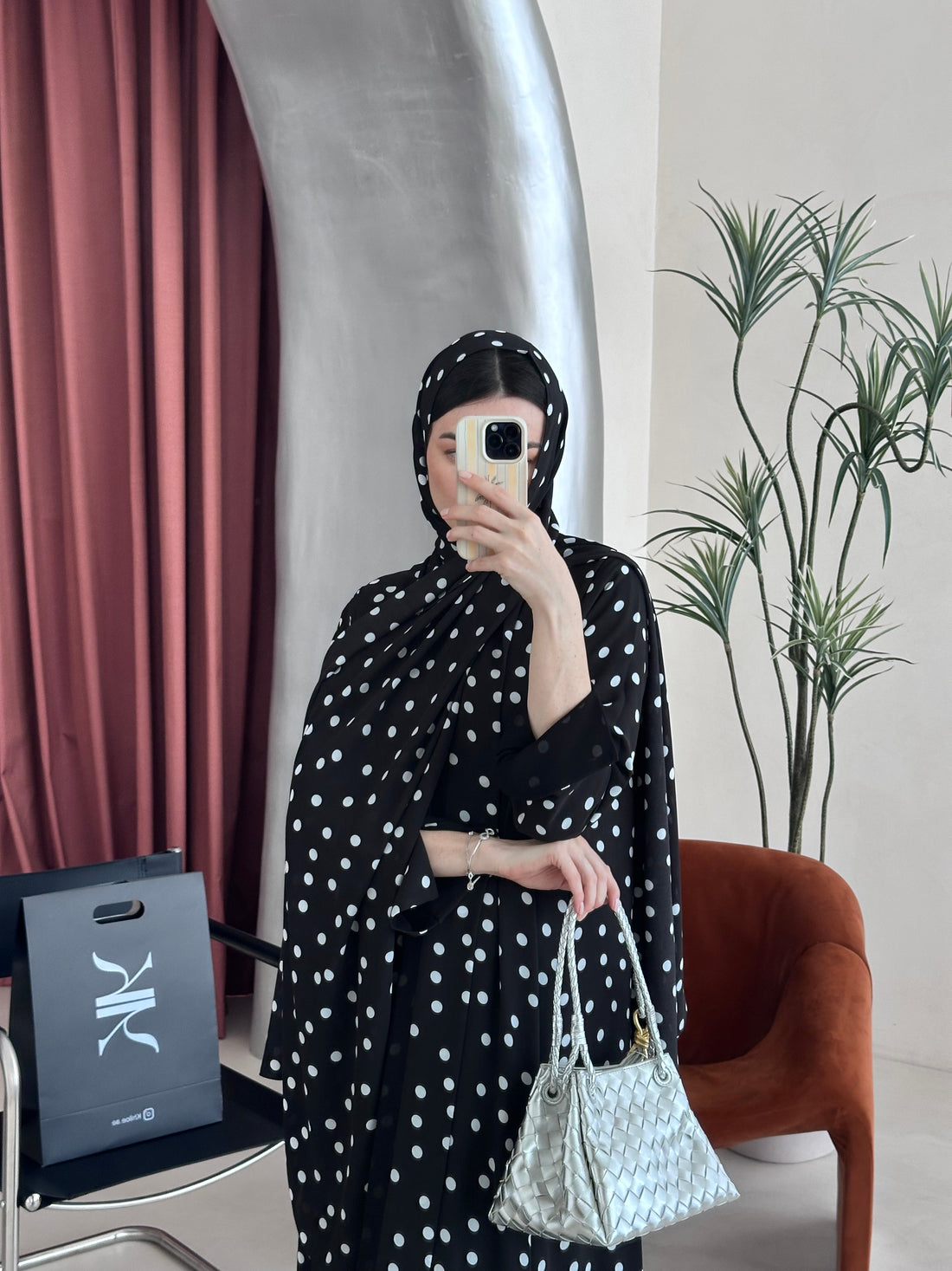 Person in a black polka dot dress holding a phone, standing next to a suitcase with a visible brand logo.