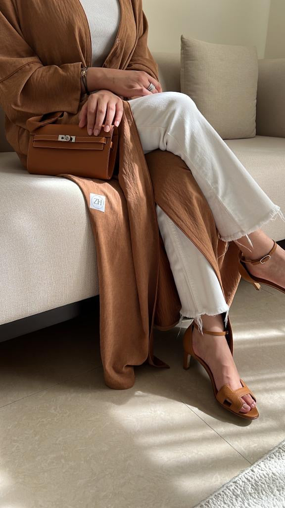 Person sitting on a couch wearing a brown coat, white pants, and brown shoes, holding a brown handbag.