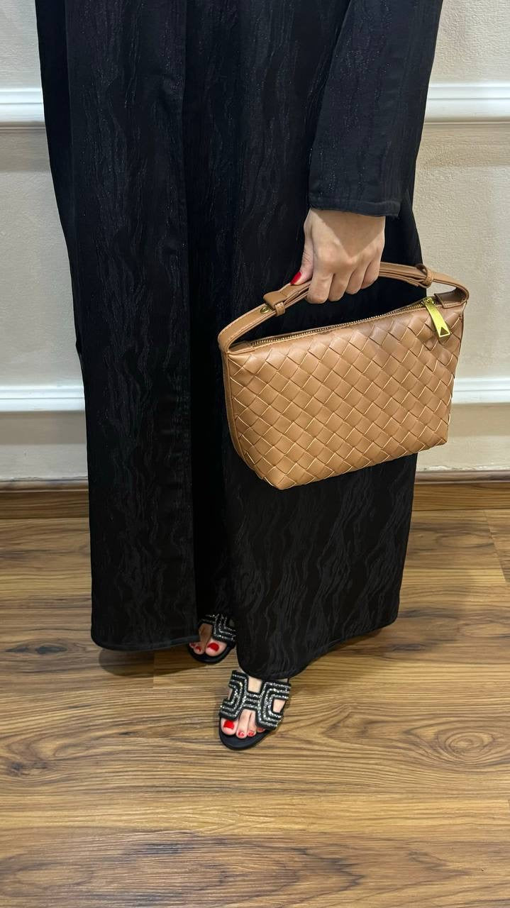 A person wearing a black abaya holding a brown, solid-patterned handbag with an adjustable shoulder strap.