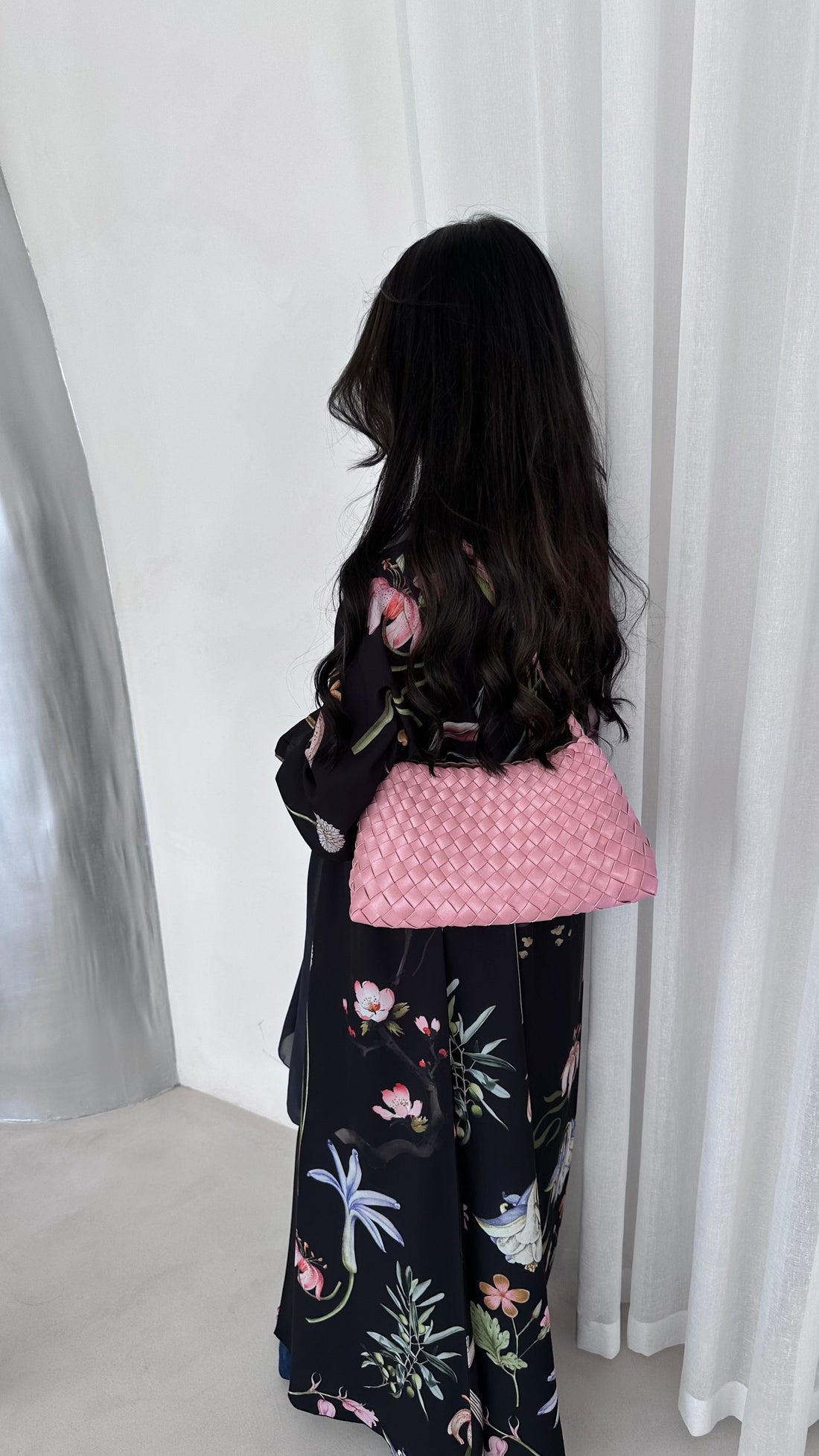 Person wearing a floral dress with a pink bag against a white curtain background