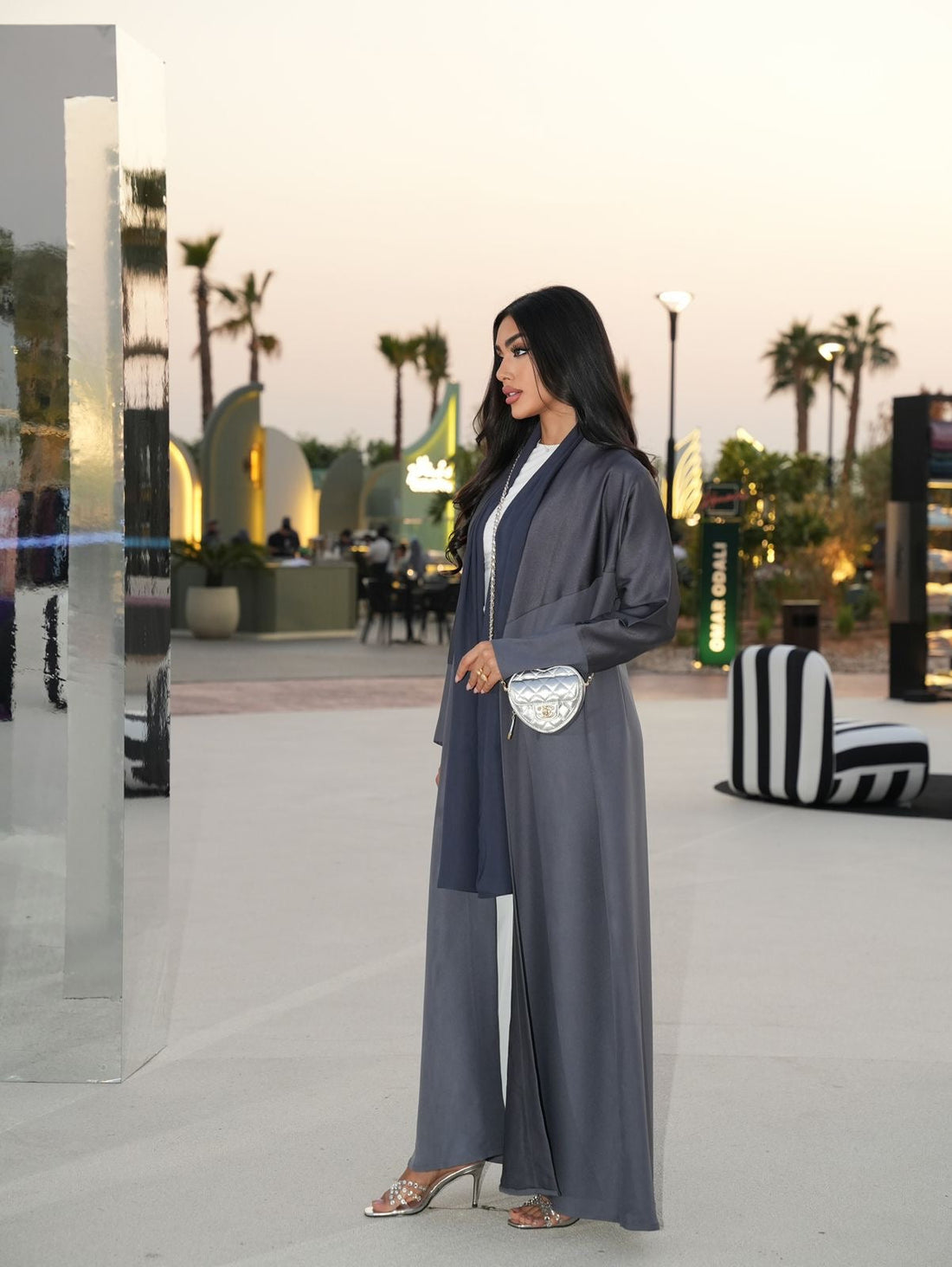 Woman in a gray outfit standing in an outdoor setting with palm trees and modern architecture.