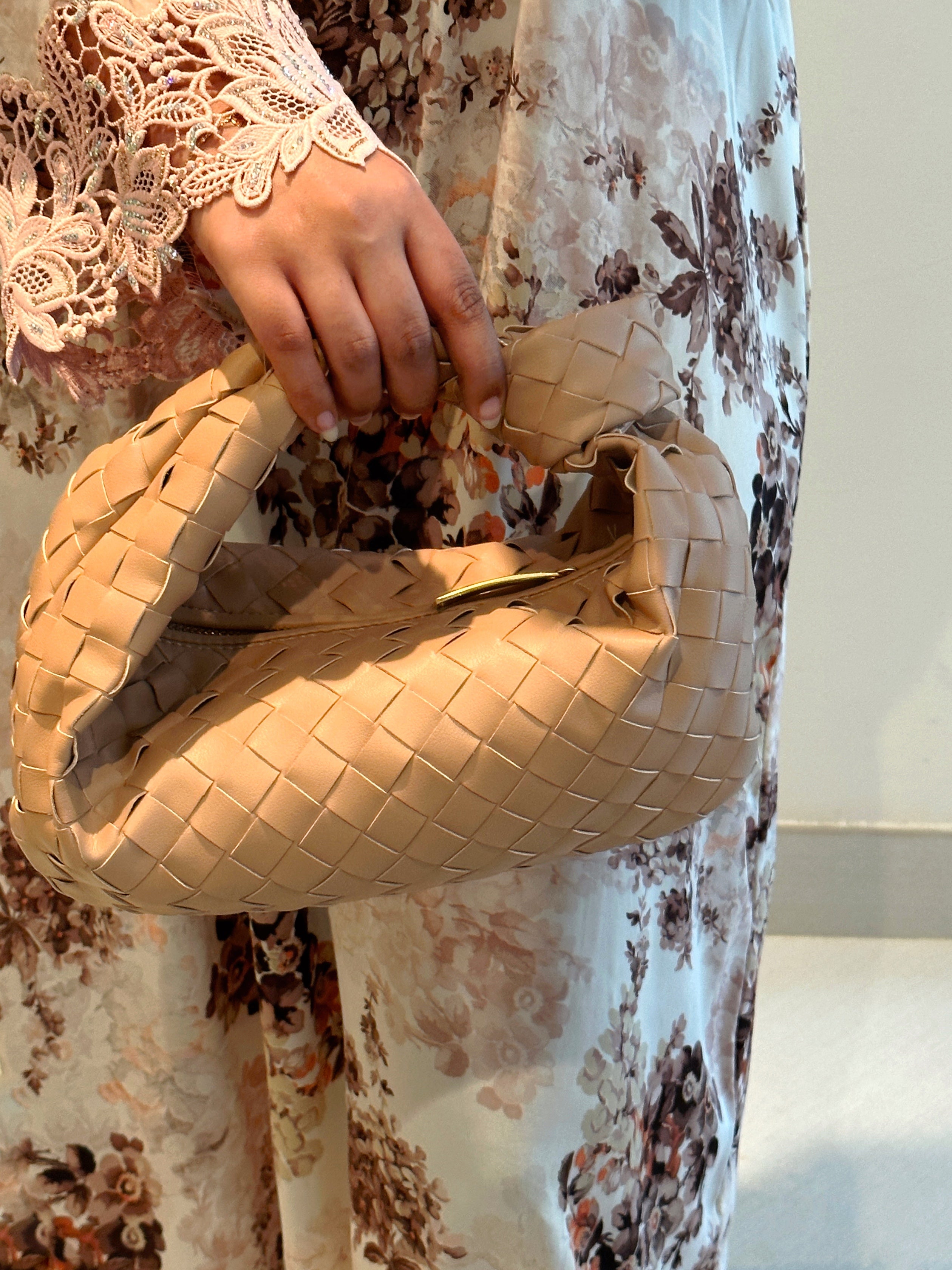 A person holding a beige leather shoulder bag with intricate weaving, fitting an iPhone pro max, and the bag is shown in a floral patterned background.