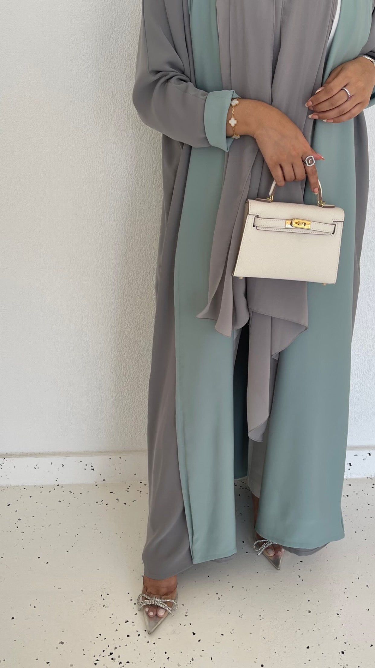 A person holding a white handbag with a capacity of 19cm, wearing a grey abaya and carrying high heels on their feet.