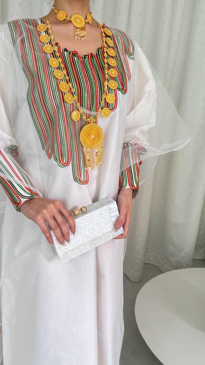 Woman wearing a white dress with colorful embroidery and yellow accessories against a white curtain.
