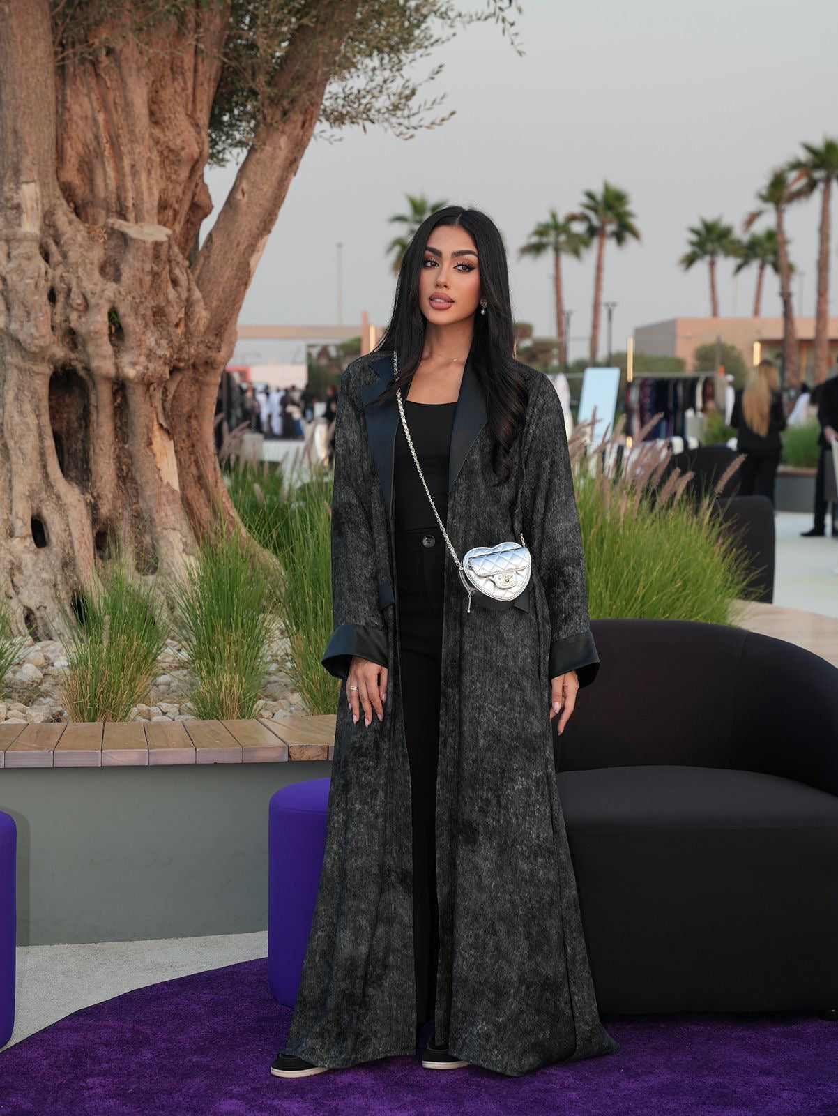 Woman in a long dark coat standing in an outdoor setting with palm trees and a purple chair.