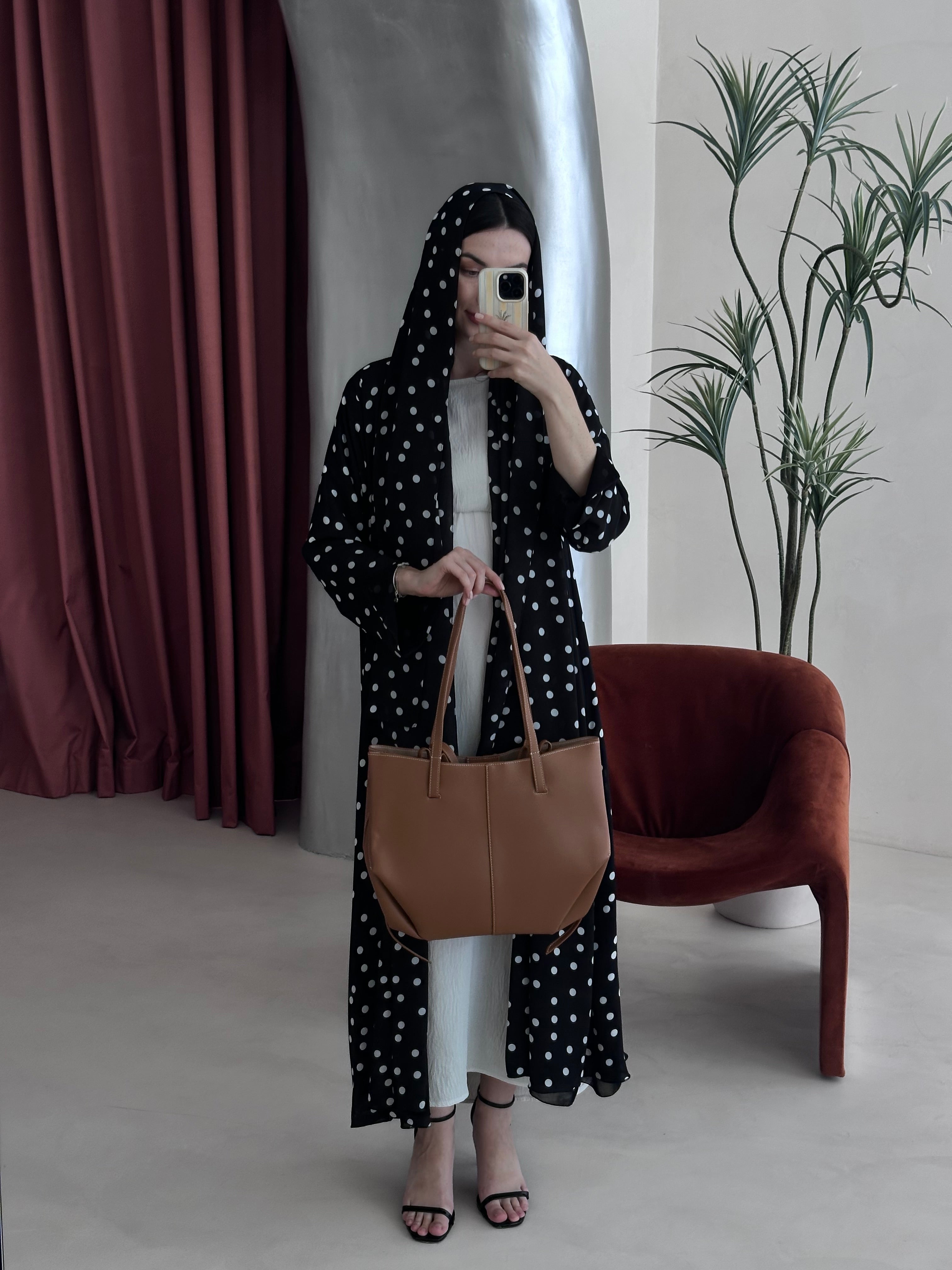 Person wearing a black polka dot dress with a brown bag in a minimalistic room.