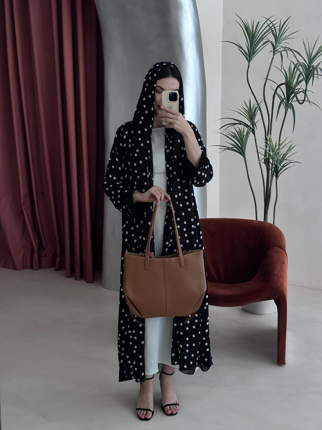 Person wearing a black polka dot dress with a brown bag in a minimalistic room.
