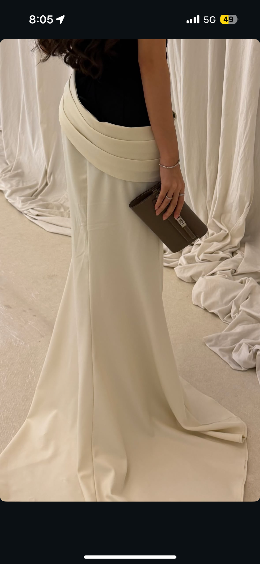 Person wearing a long, flowing white dress with a black top against a draped white curtain background.