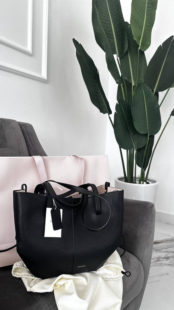 A black mini tote bag placed on a chair with a larger bag and a plant in the background.