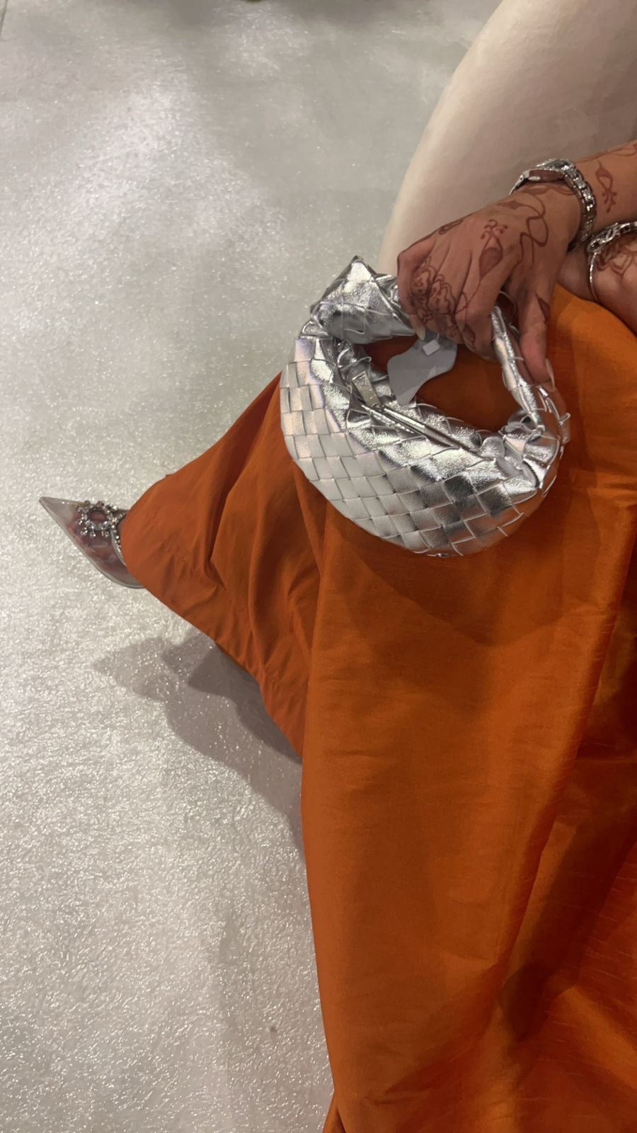 Person wearing a silver quilted handbag with an orange garment and light gray floor.