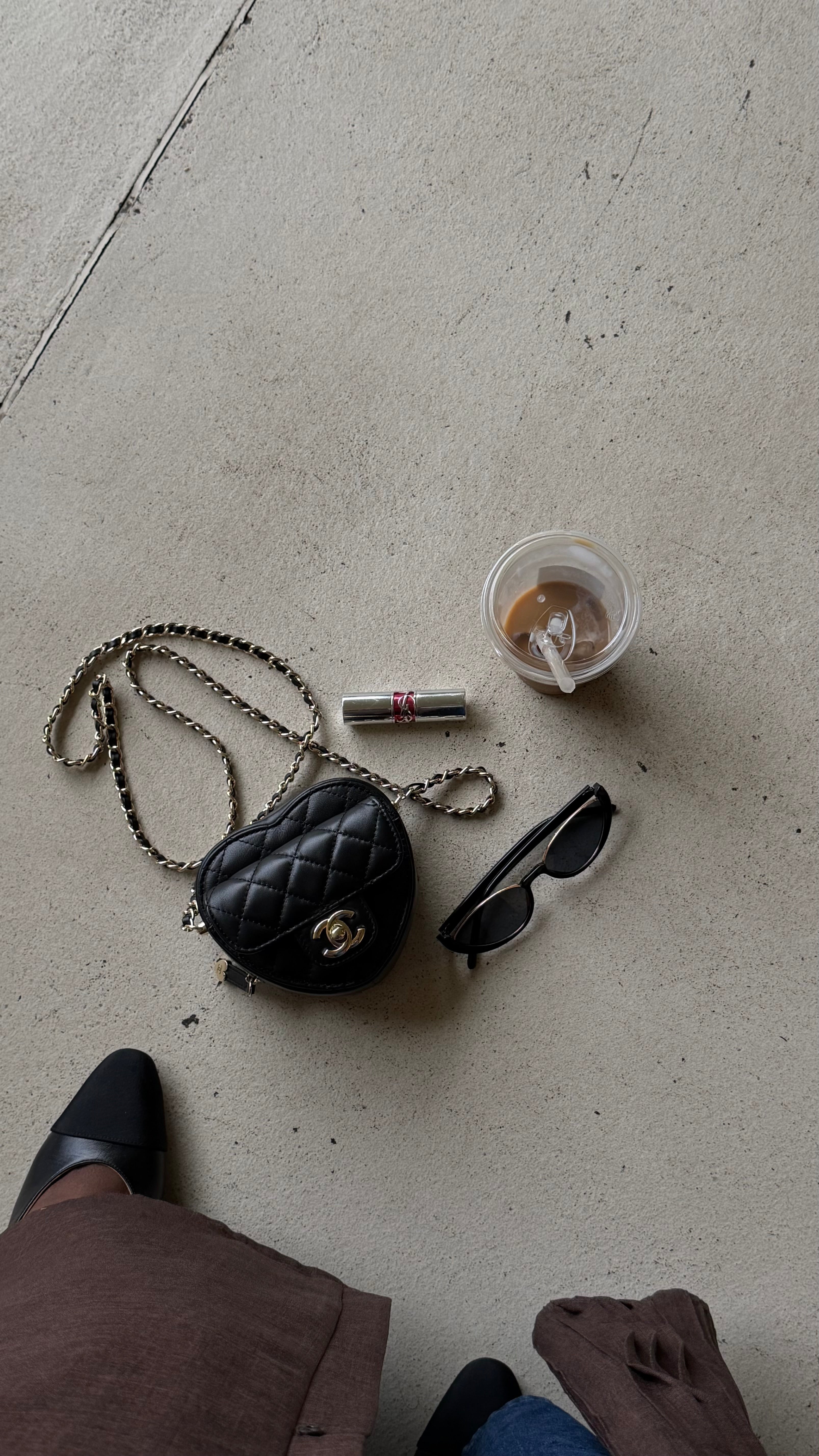Black handbag, sunglasses, and a small container on a concrete floor with a person's feet visible.