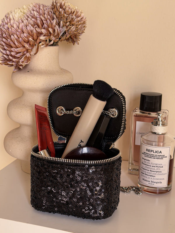 Black sequined makeup bag with cosmetic items on a surface with a decorative vase in the background.
