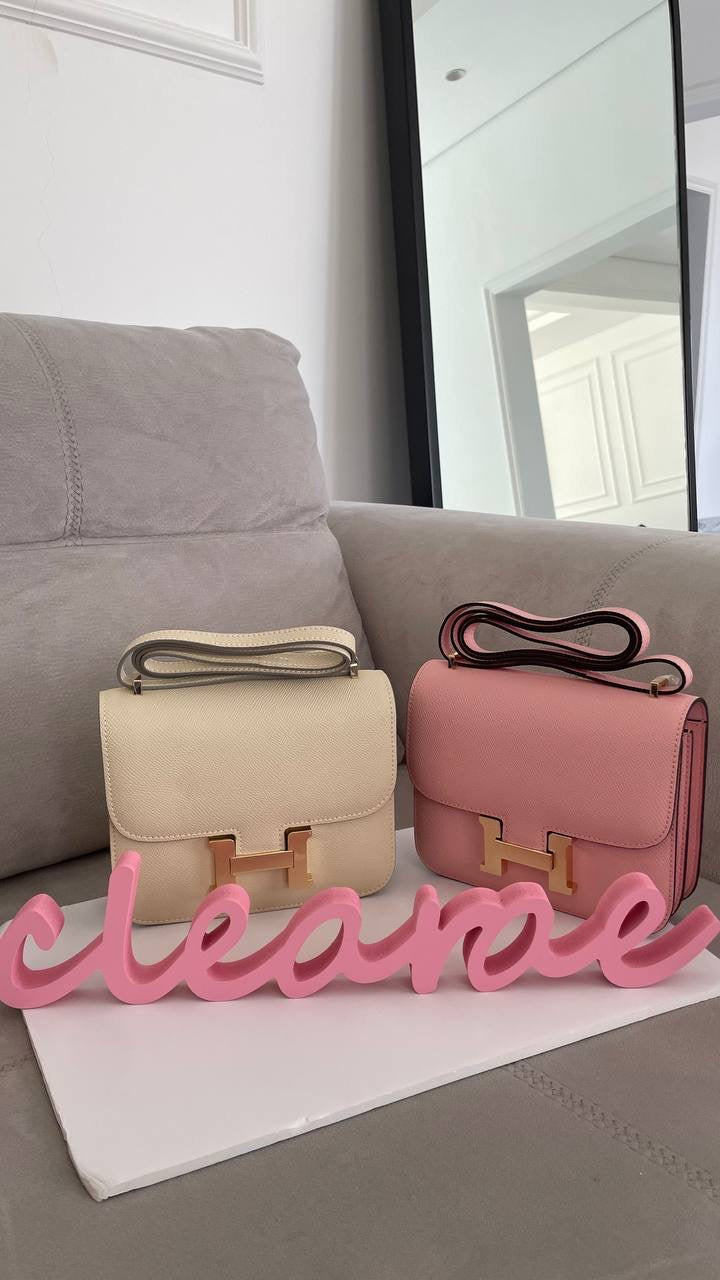 Two handbags, one beige and one pink, on a surface with a decorative sign reading 'cleanse' in front of a gray couch.