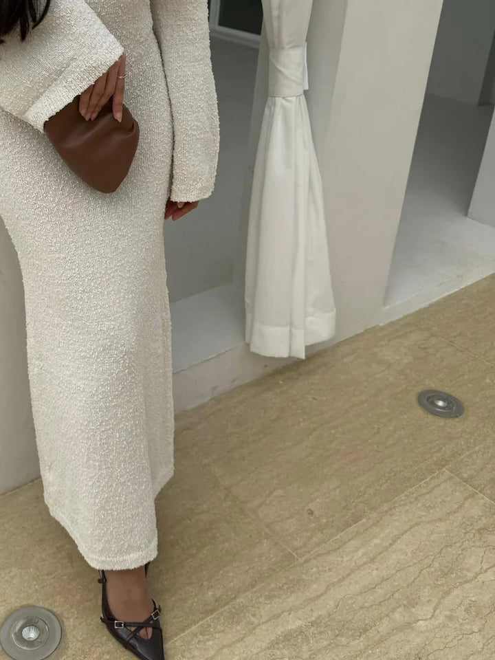 Person wearing a textured white coat and pants, holding a brown handbag, standing on a wooden floor.