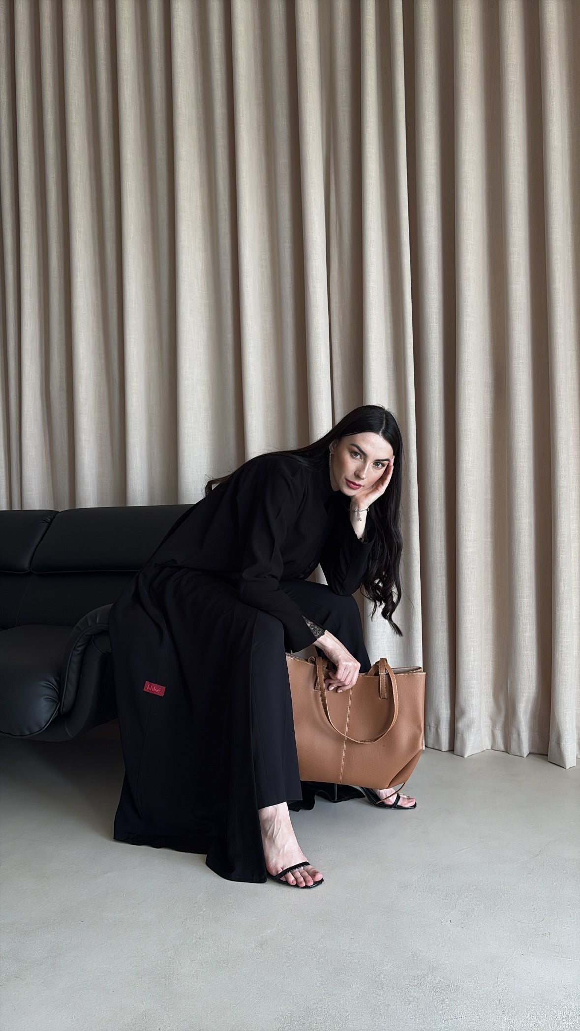 Woman in black outfit holding a brown leather bag against a beige curtain background