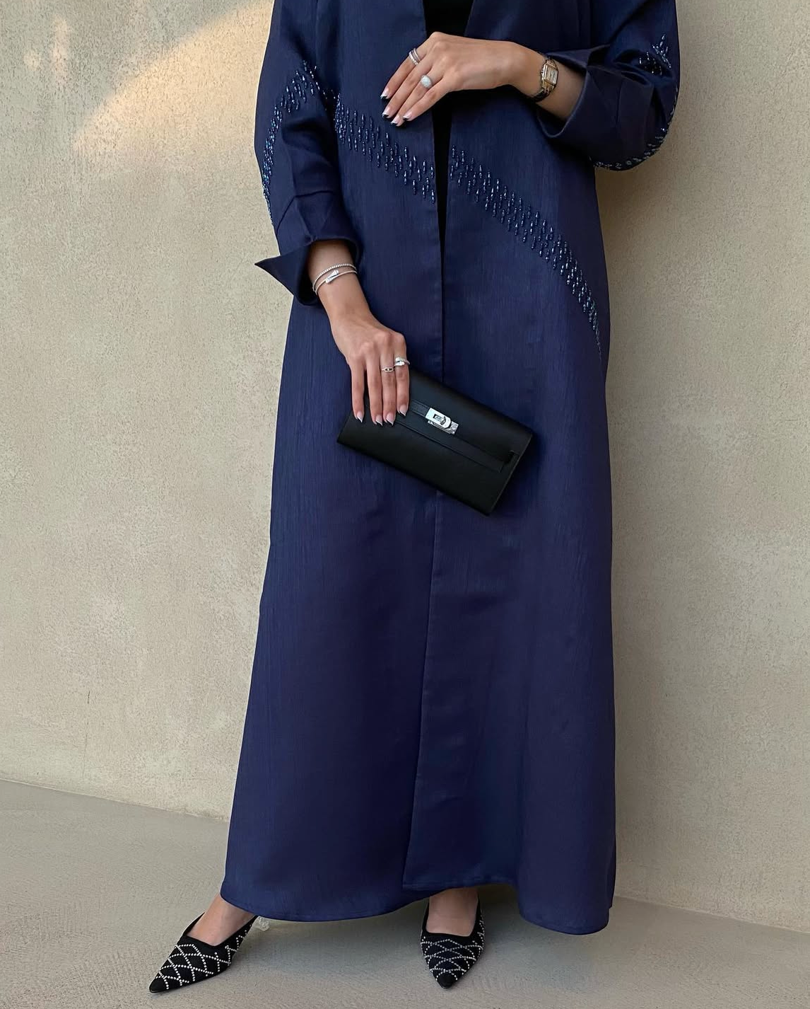 Person wearing a long navy blue dress with intricate detailing, holding a black clutch against a neutral background.