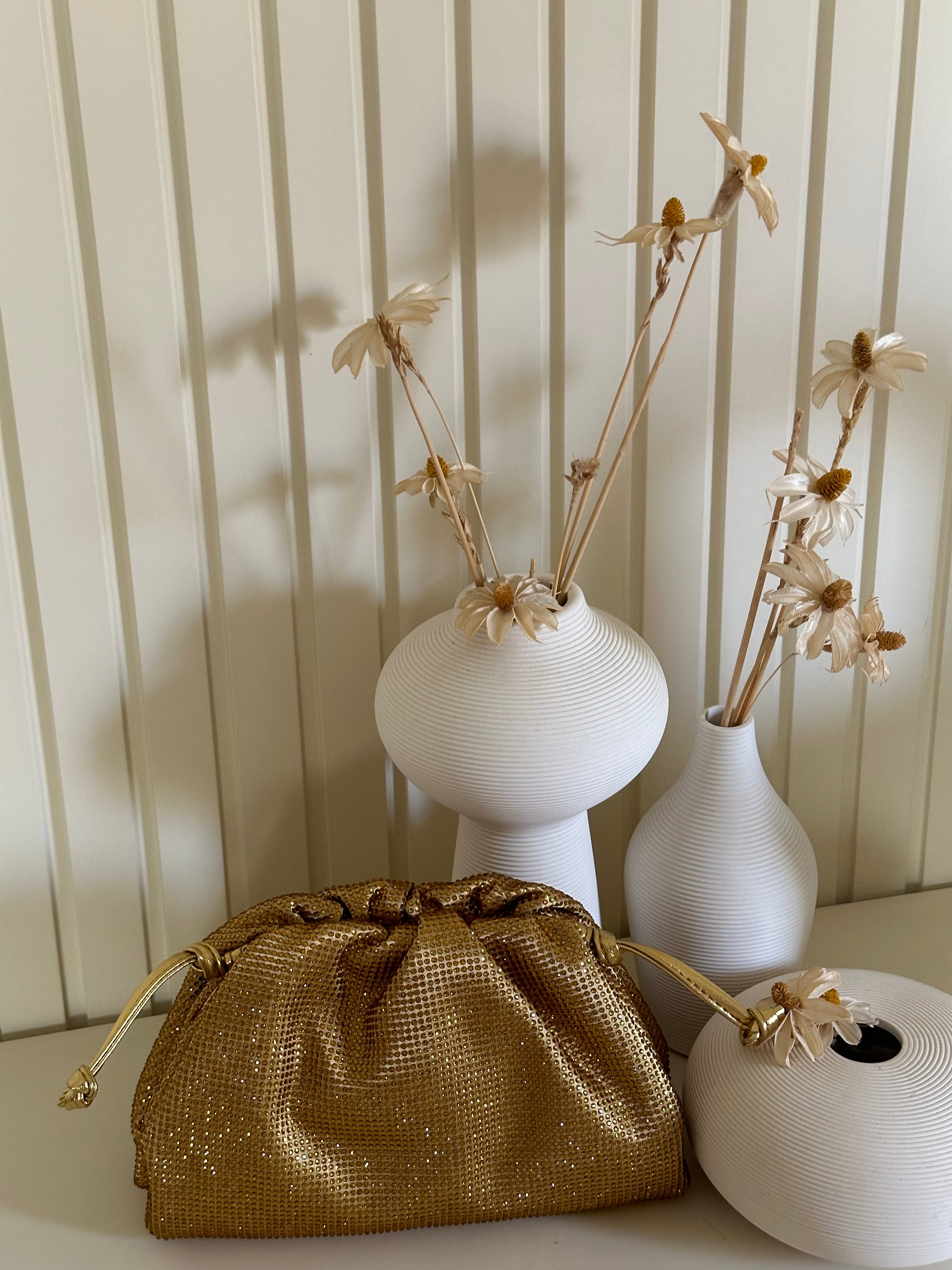 Gold drawstring bag with white vases and dried flowers against a striped wall.