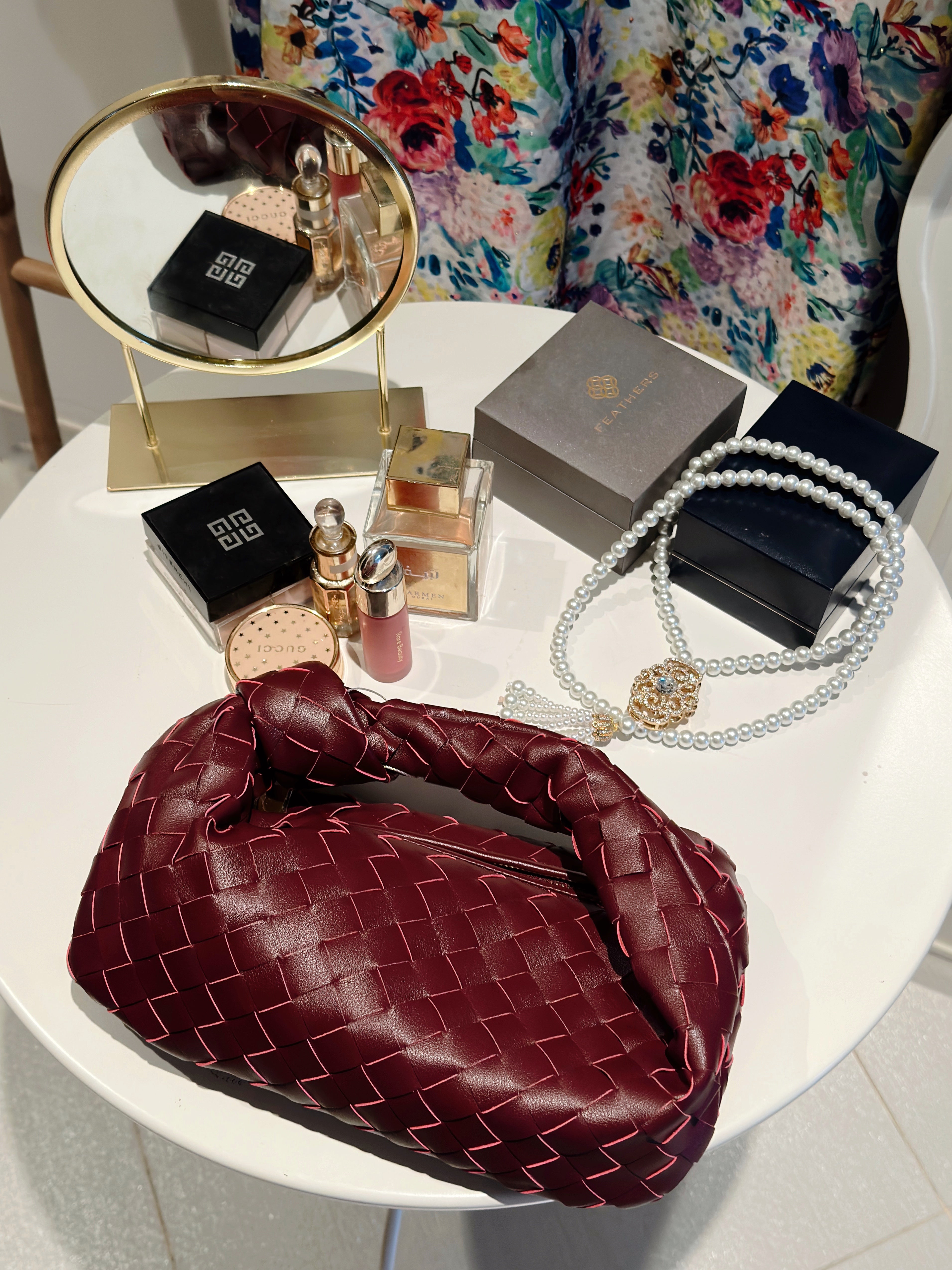 Red intrecciato leather shoulder bag displayed on a white surface, accompanied by various accessories such as a mirror, boxes, and jewelry.
