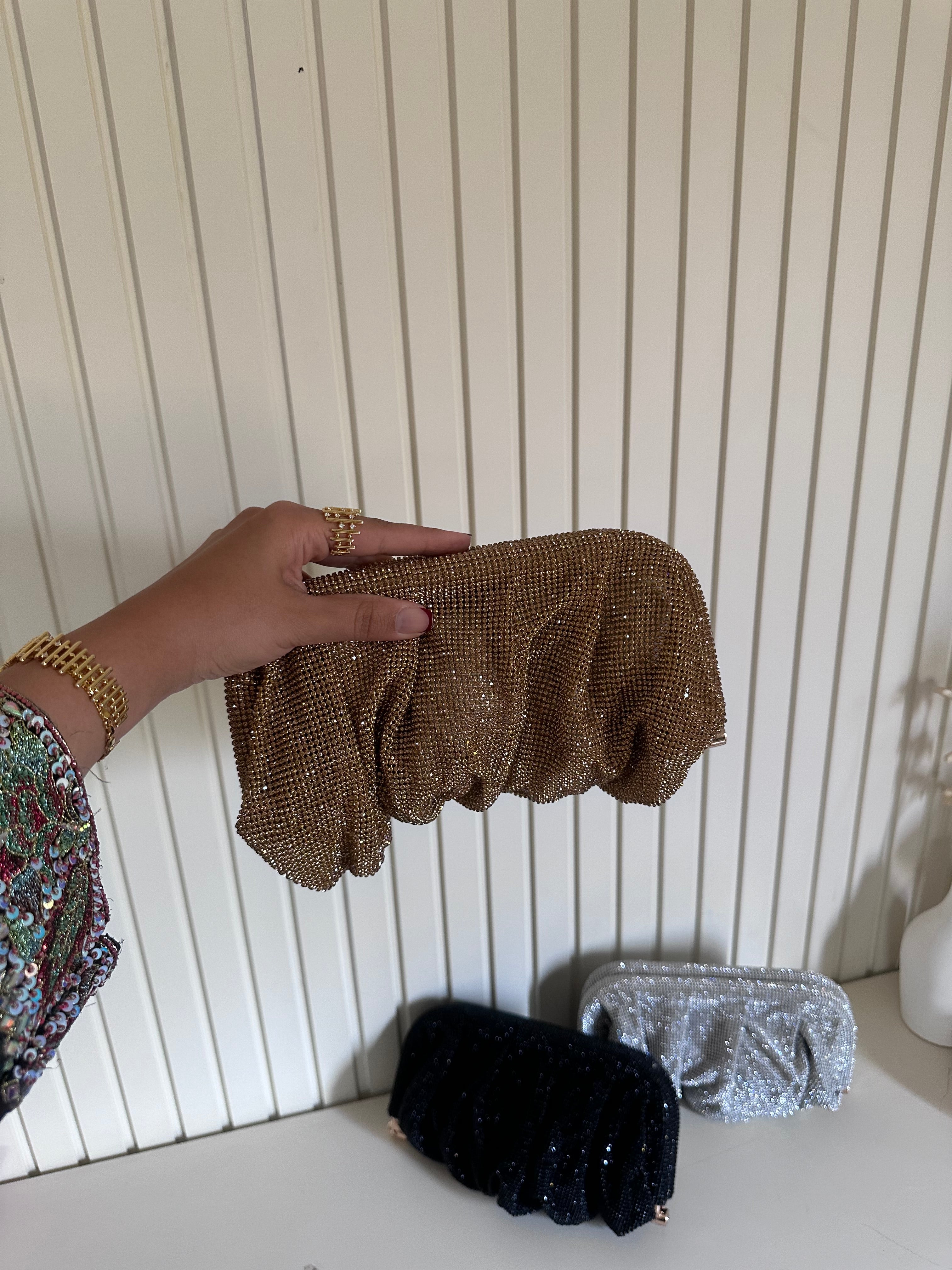 Hand holding a gold clutch with other clutches on a light wooden surface.