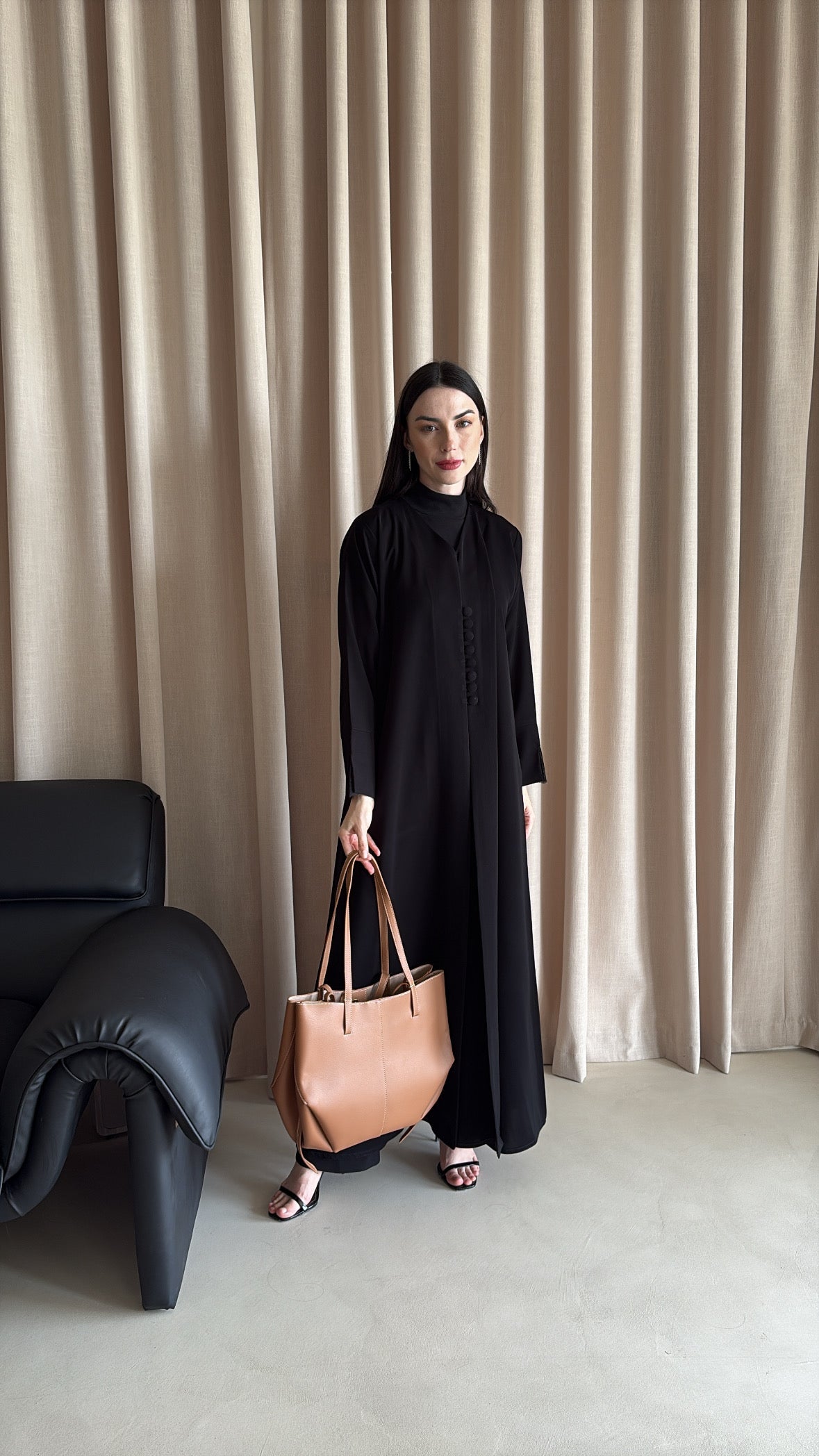 Woman in a black outfit holding a brown bag against a beige curtain background