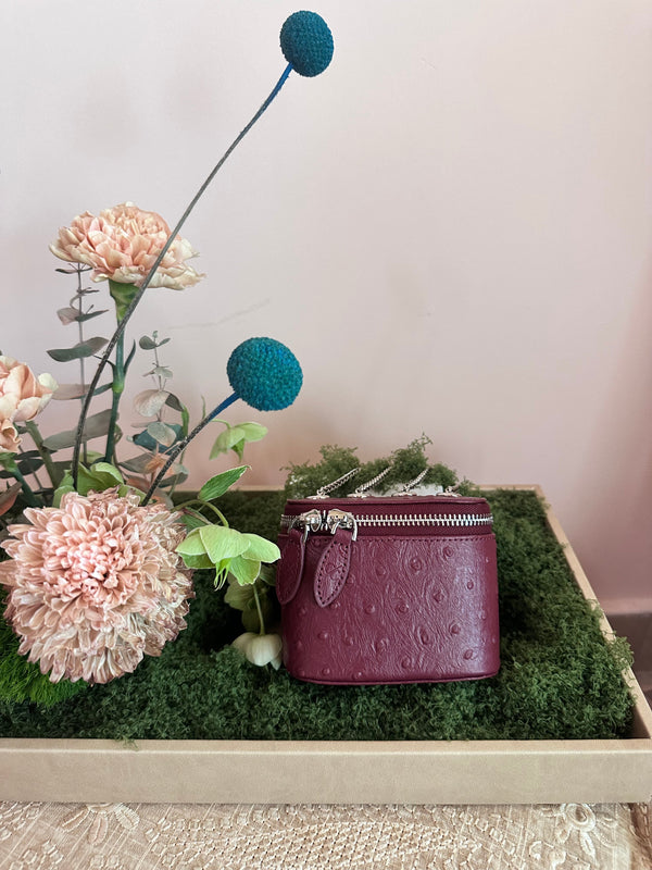 Maroon handbag on a bed of green moss with decorative flowers and plants.