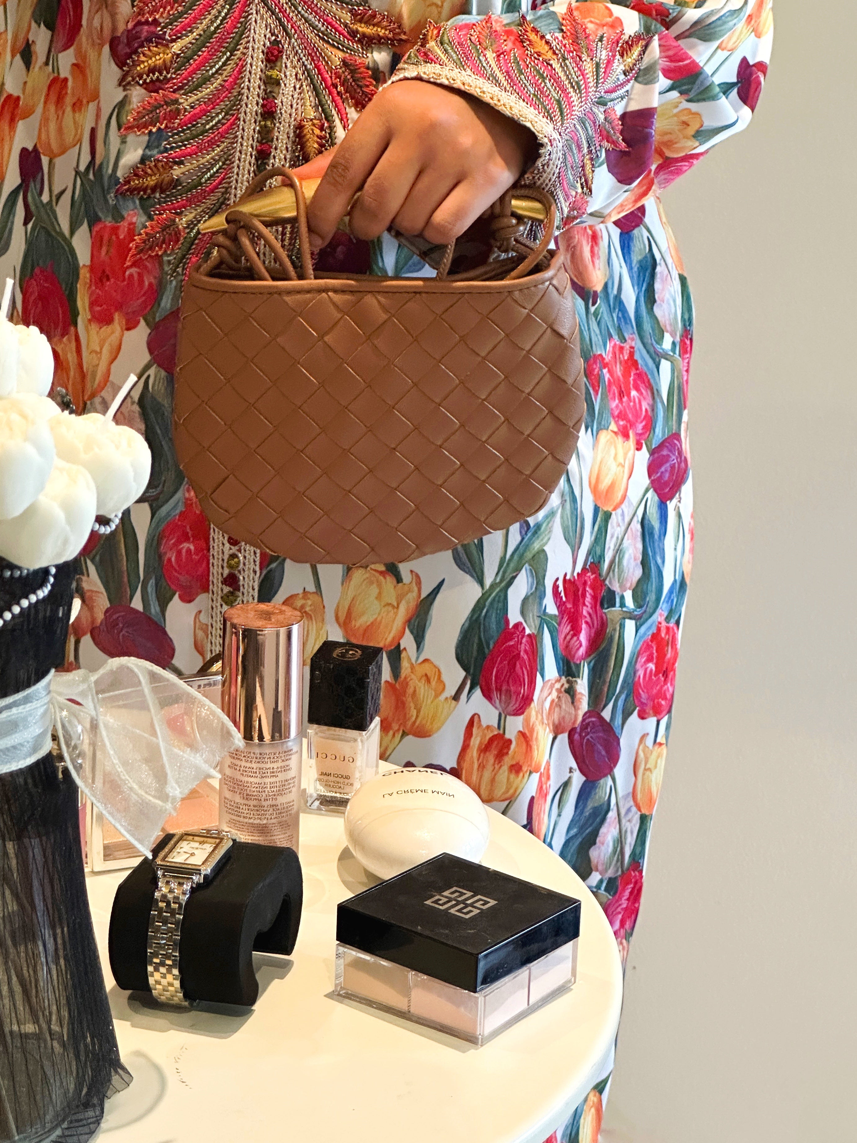 A person holding a brown mini bag with a quilted design, in front of a display that includes cosmetics and other accessories.