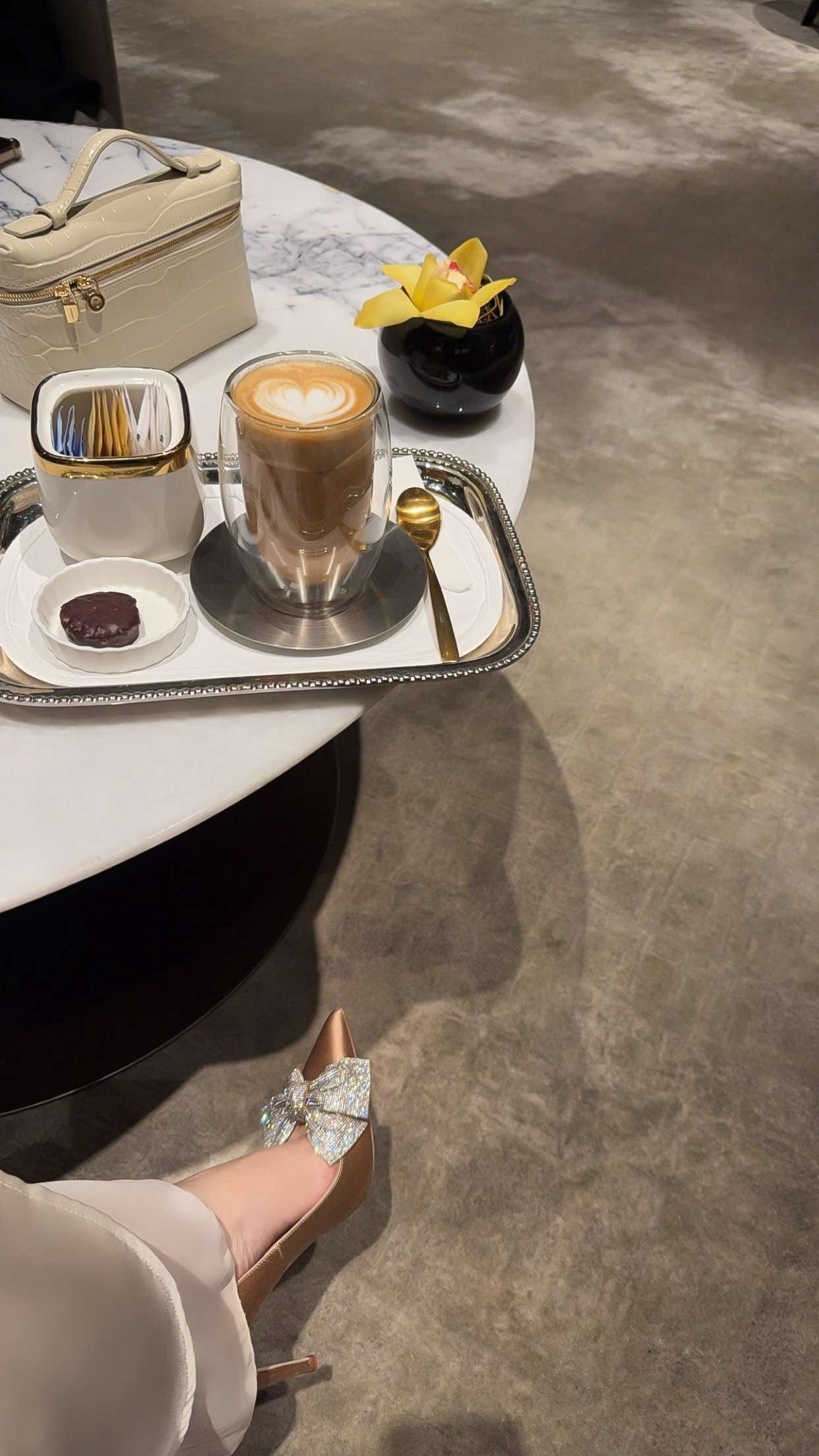 Café setting with a glass of coffee on a marble table, a hand reaching out, and a handbag.