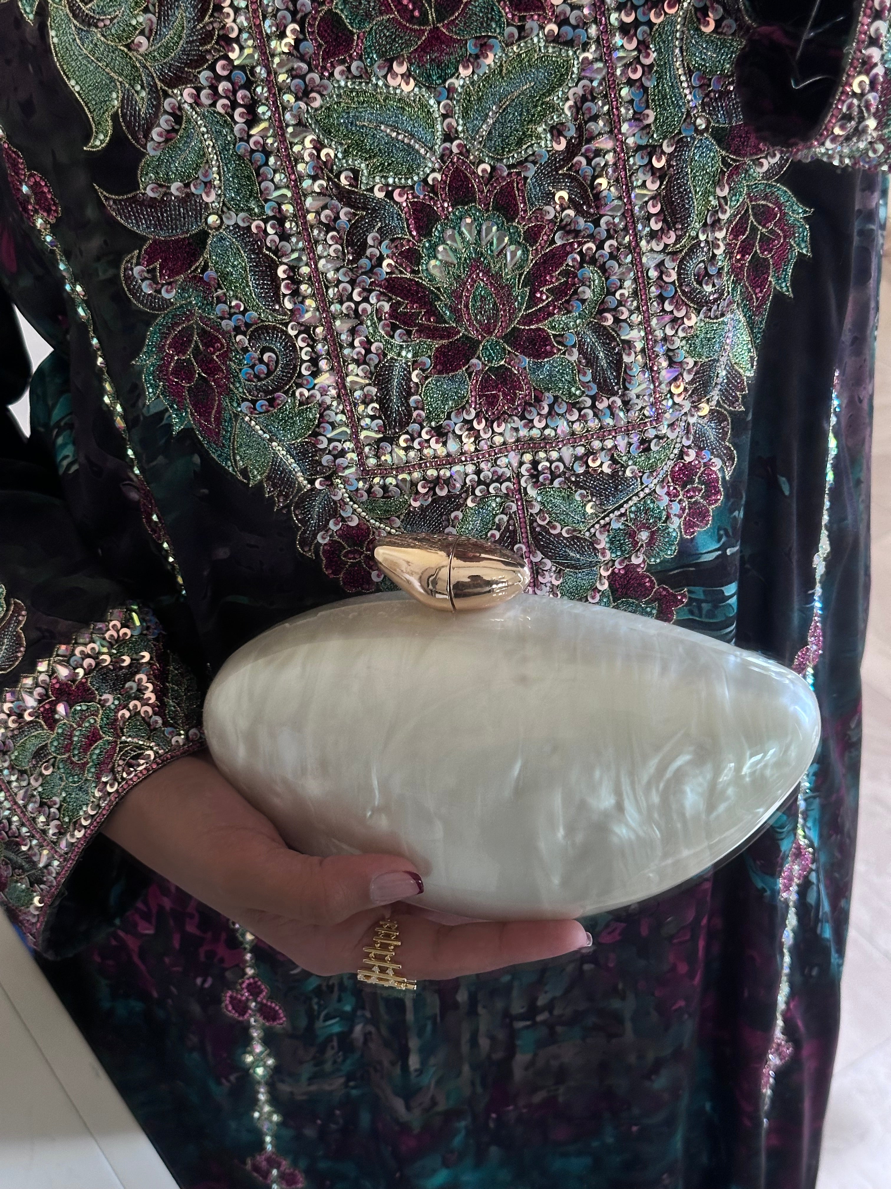 Hand holding a white clutch with a person wearing a dark, intricately embroidered garment.