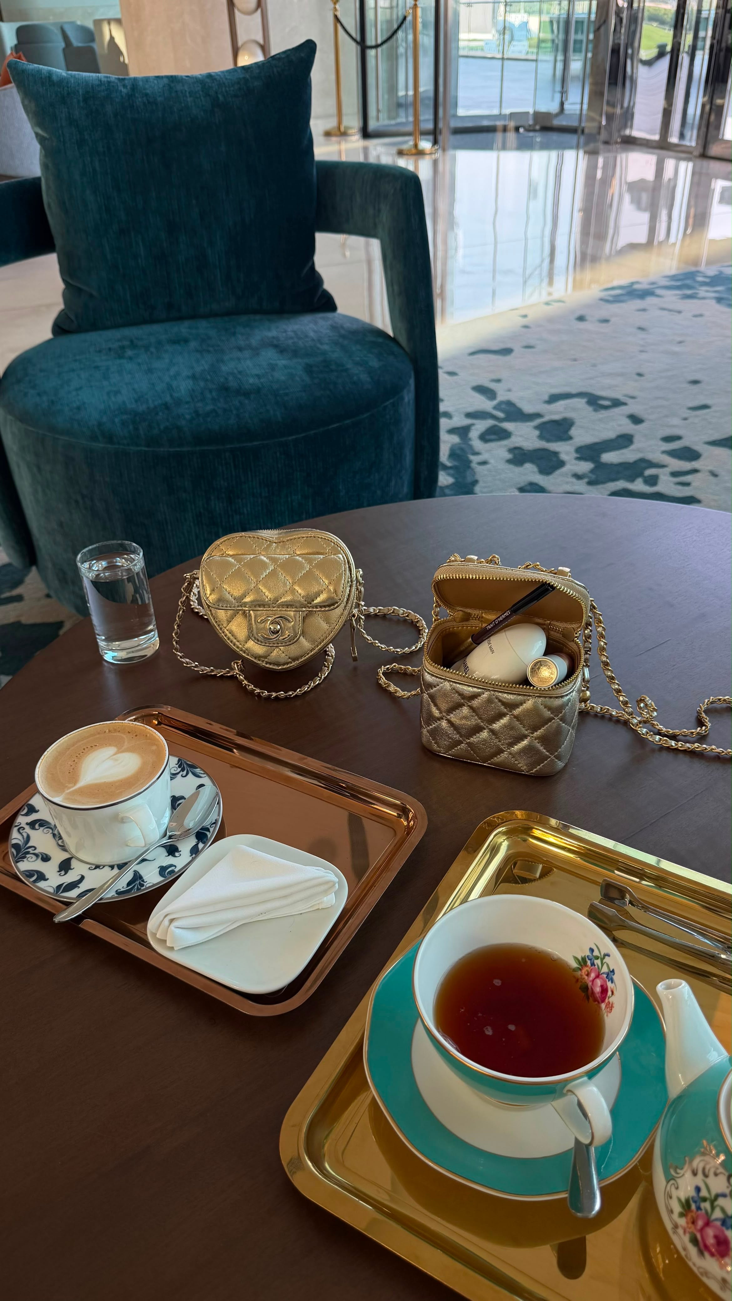 A styled cafe table setting featuring a quilted gold shoulder bag, a small quilted beige bag with a chain strap, and teacups on trays in a modern lounge setting.