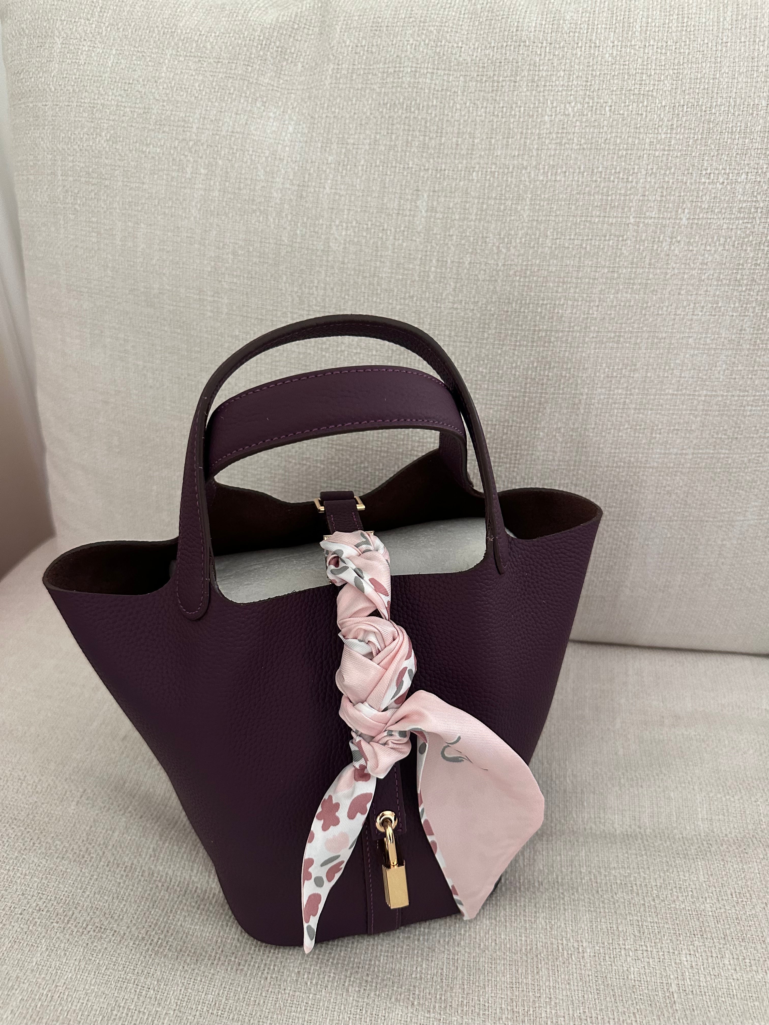 A purple handbag with a soft, foldable texture, featuring a key for a lock and a pink scarf tied to its handle.