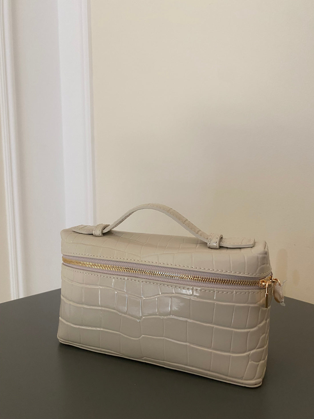 Beige crocodile-patterned handbag with a gold zipper on a dark surface.
