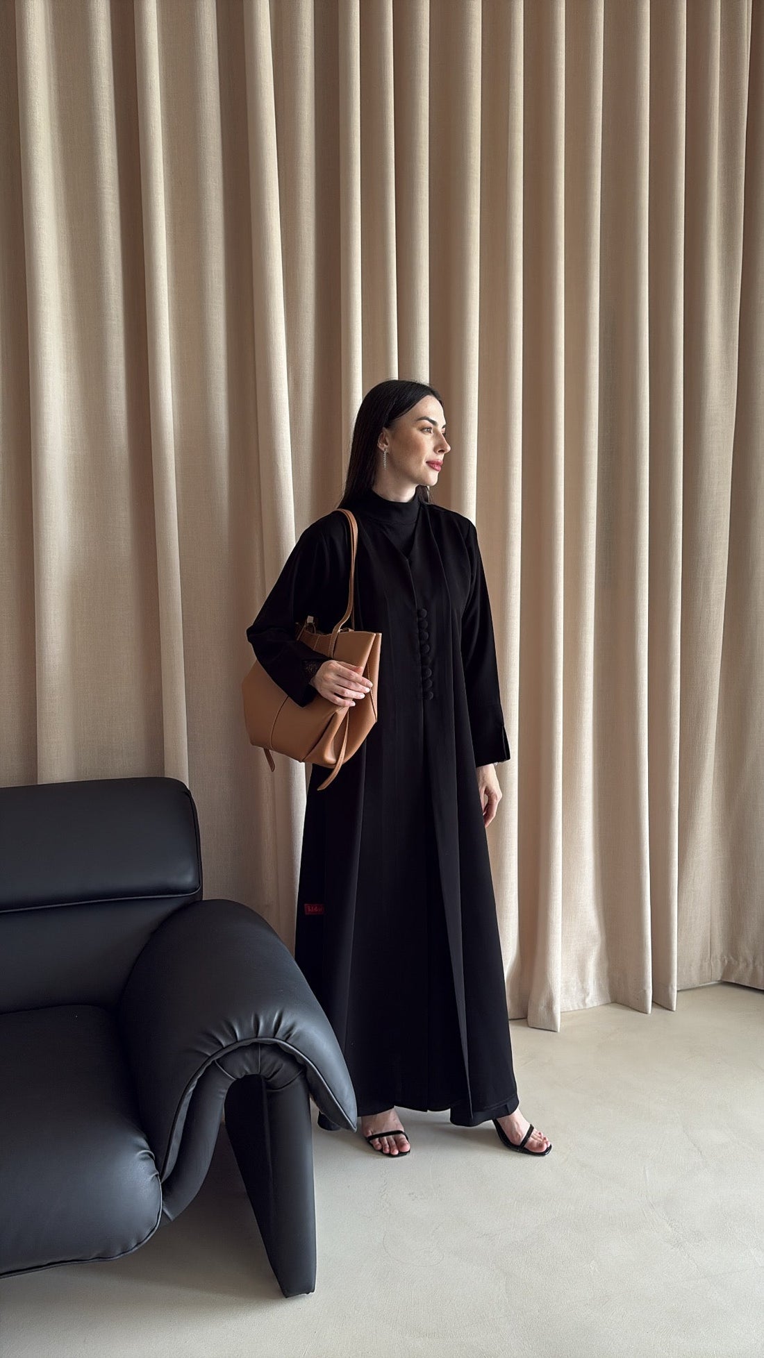 Woman in a black dress standing in a room with beige curtains and a blue sofa.