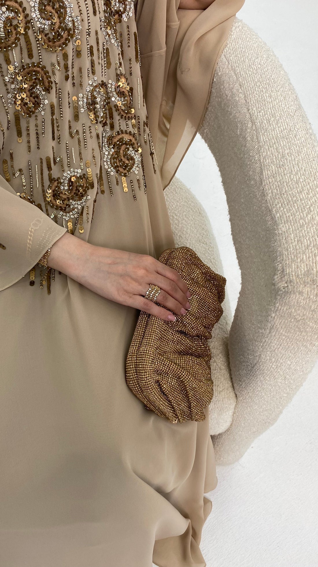 Person wearing a beige dress with gold embellishments holding a matching clutch against a white background