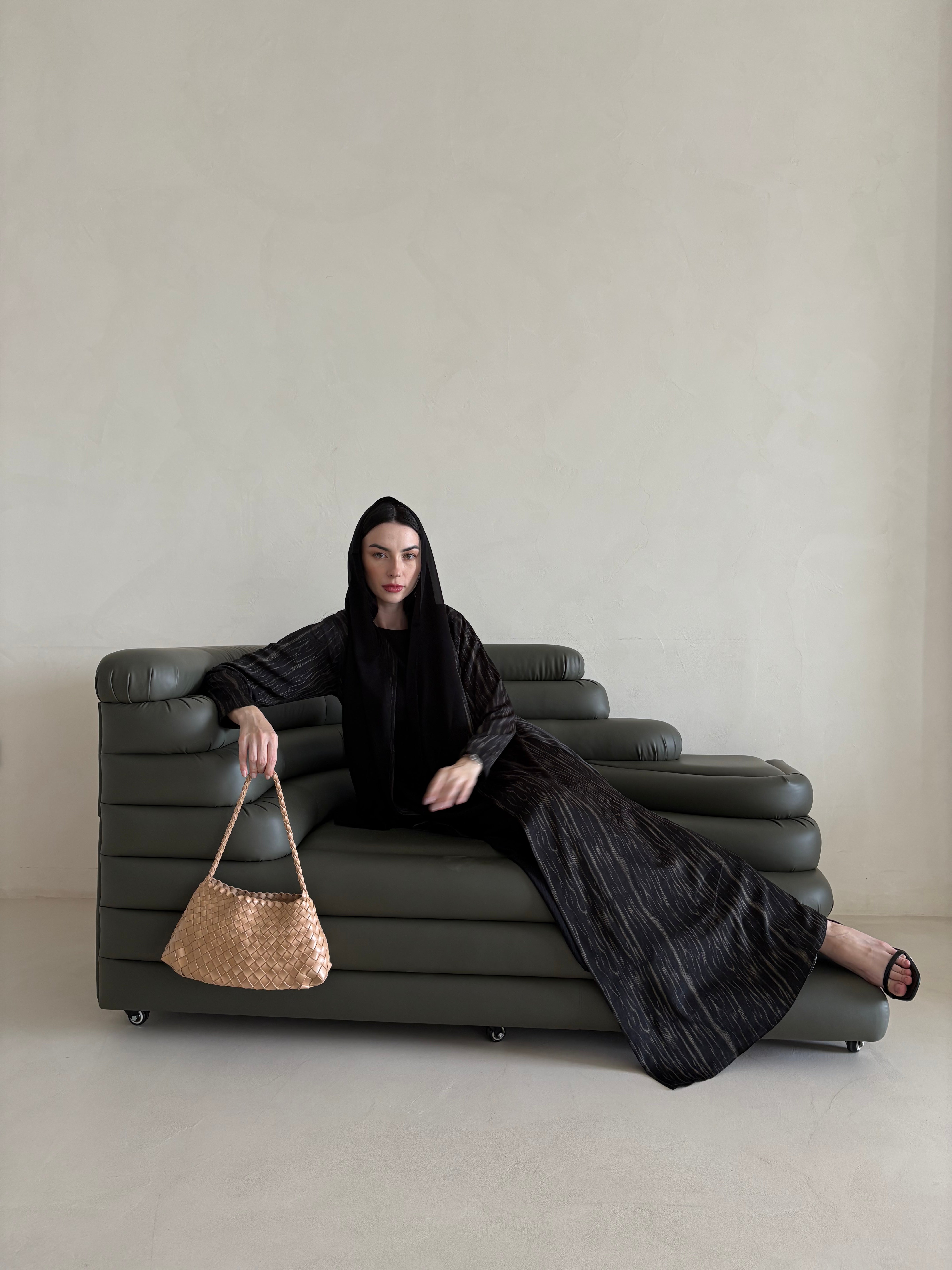 Woman in a black outfit sitting on a gray sofa holding a woven bag.