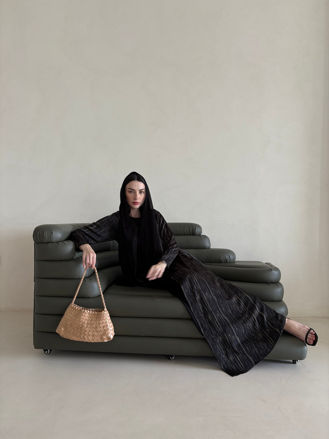 Woman in a black outfit sitting on a gray sofa holding a woven bag.