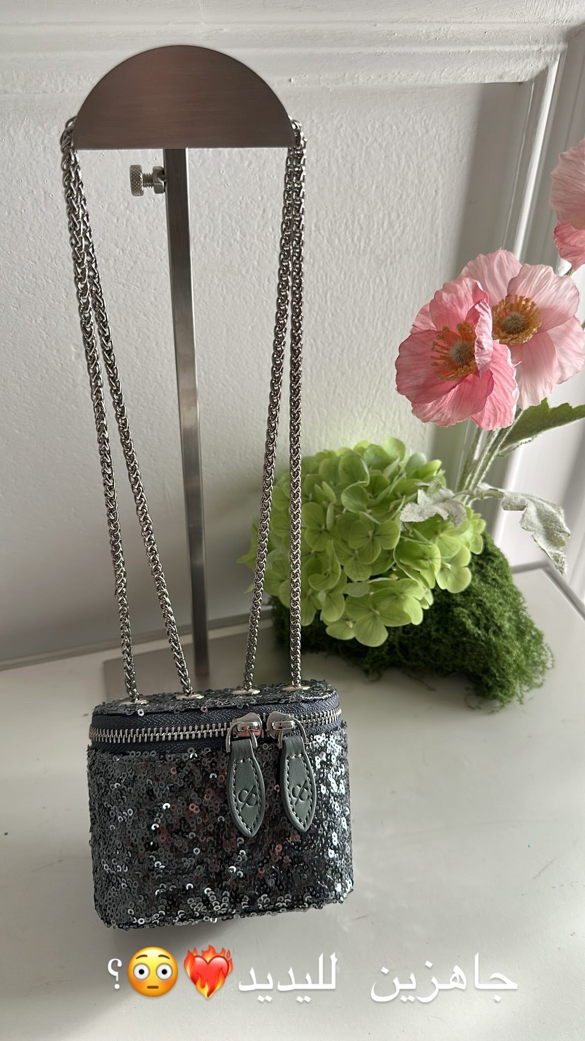 Black sequined handbag with chain strap on a stand, surrounded by flowers and greenery.
