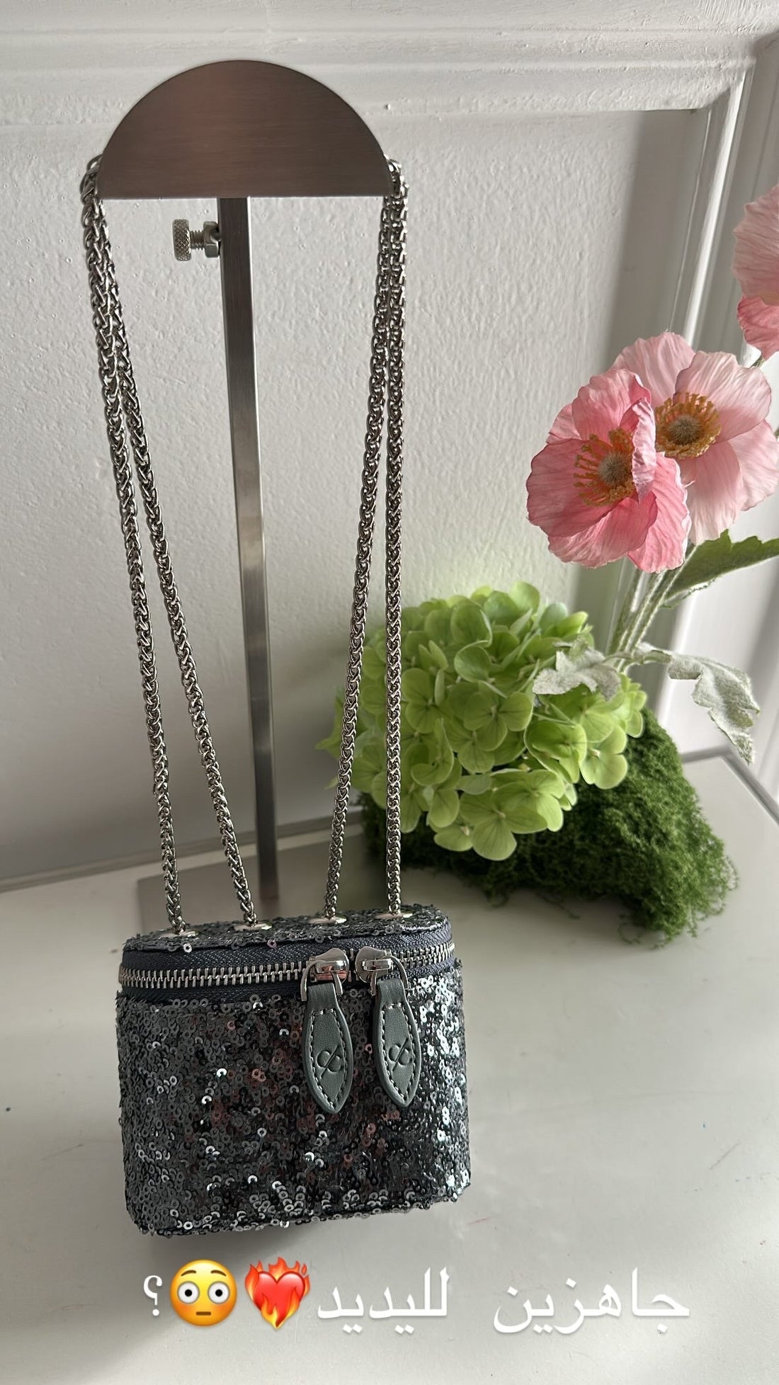 Black sequined handbag with chain strap on a stand, surrounded by flowers and greenery.