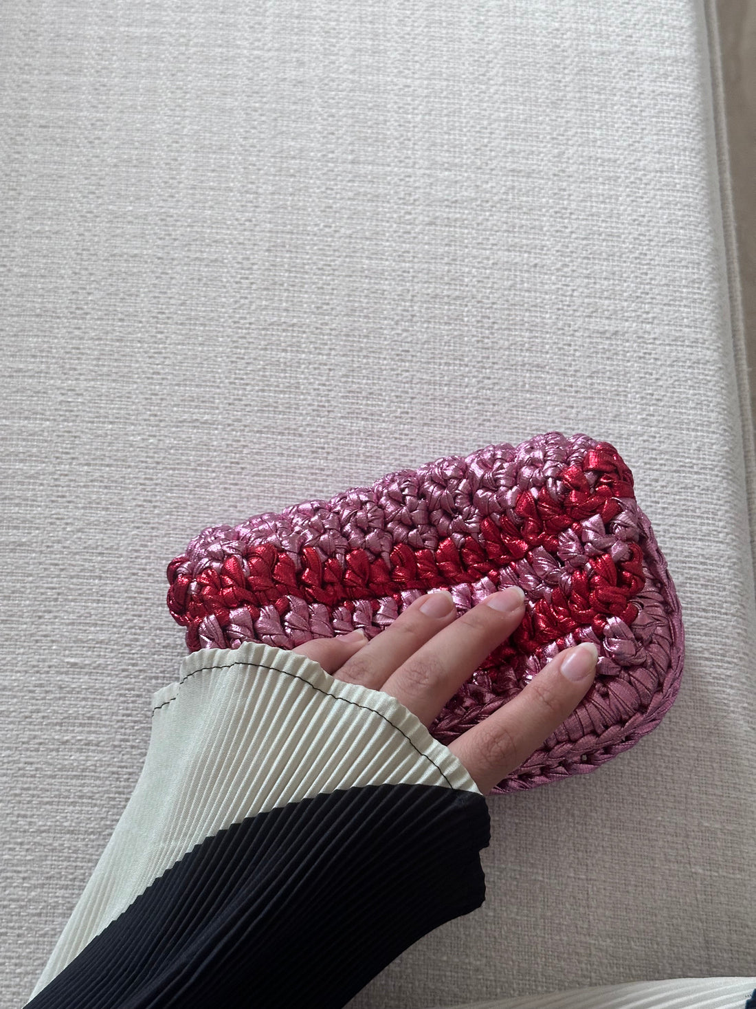 Pink and red textured clutch bag on a light gray surface with a hand reaching towards it.