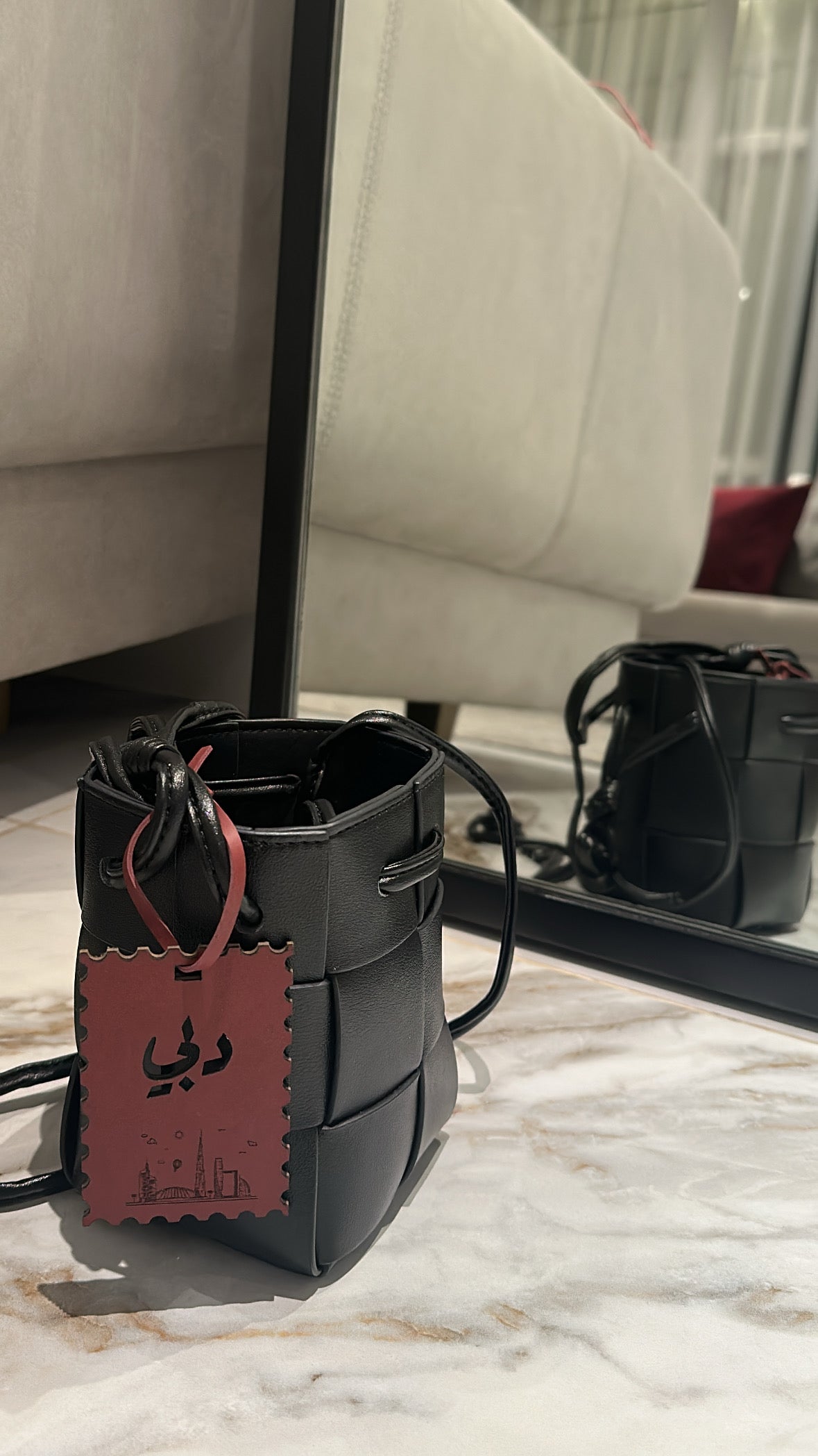 Black handbag with a red tag on a marble surface, with a blurred background of a beige couch and mirror.