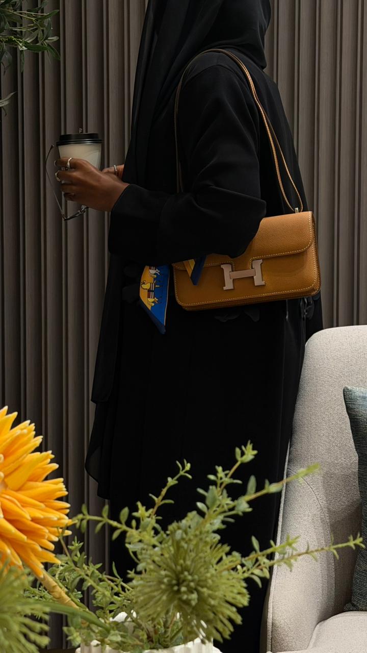 Person holding a coffee cup and a tan handbag with a visible brand logo, standing in a room with plants and a sofa.