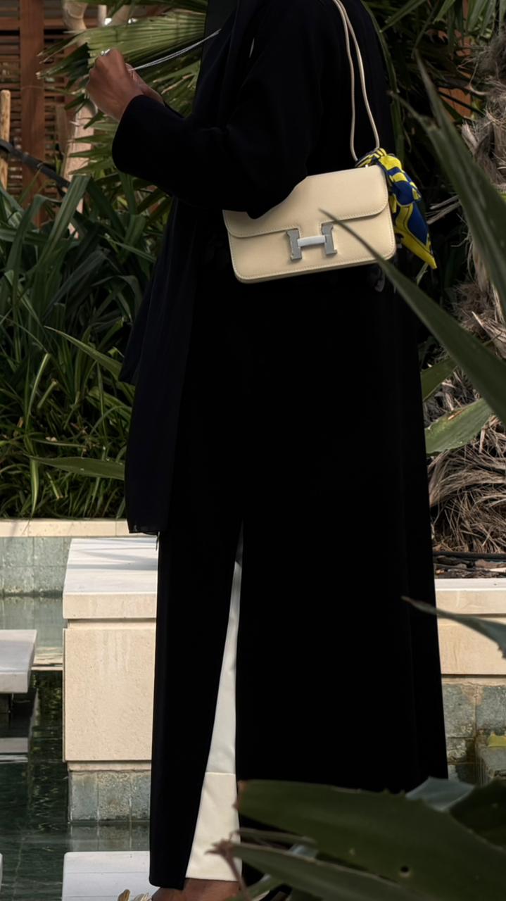 Person wearing a black outfit with a white handbag featuring a yellow 'H' logo, standing by a poolside.