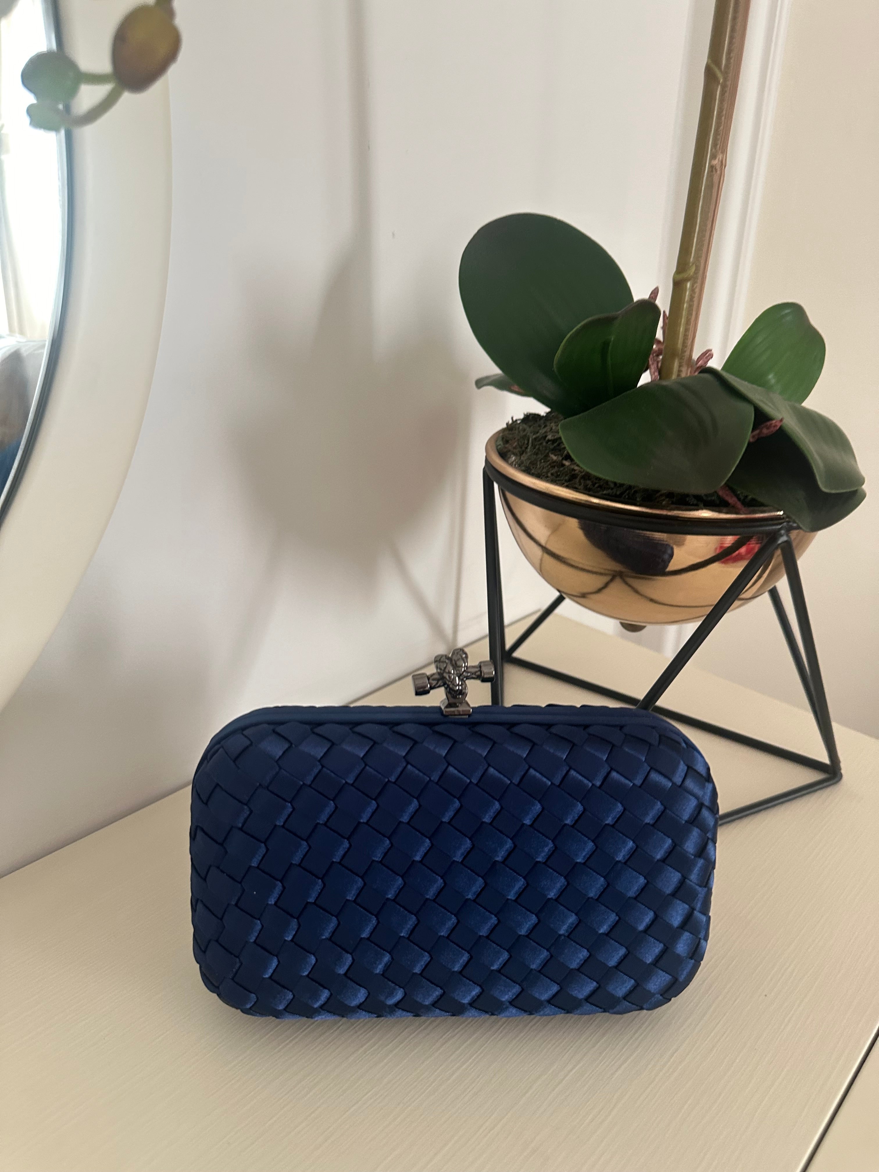 A blue clutch with a quilted design placed on a white surface, with a potted plant and a metallic object in the background.
