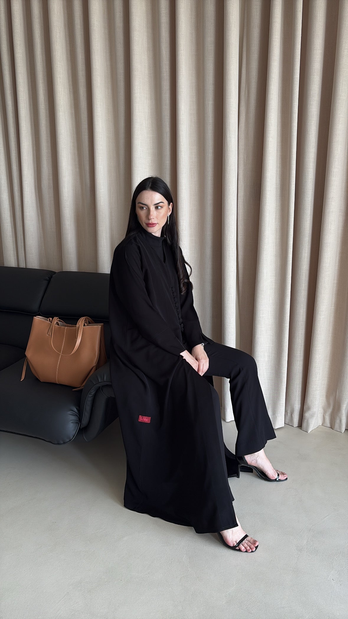 Woman in a black outfit sitting on a couch with a beige curtain background