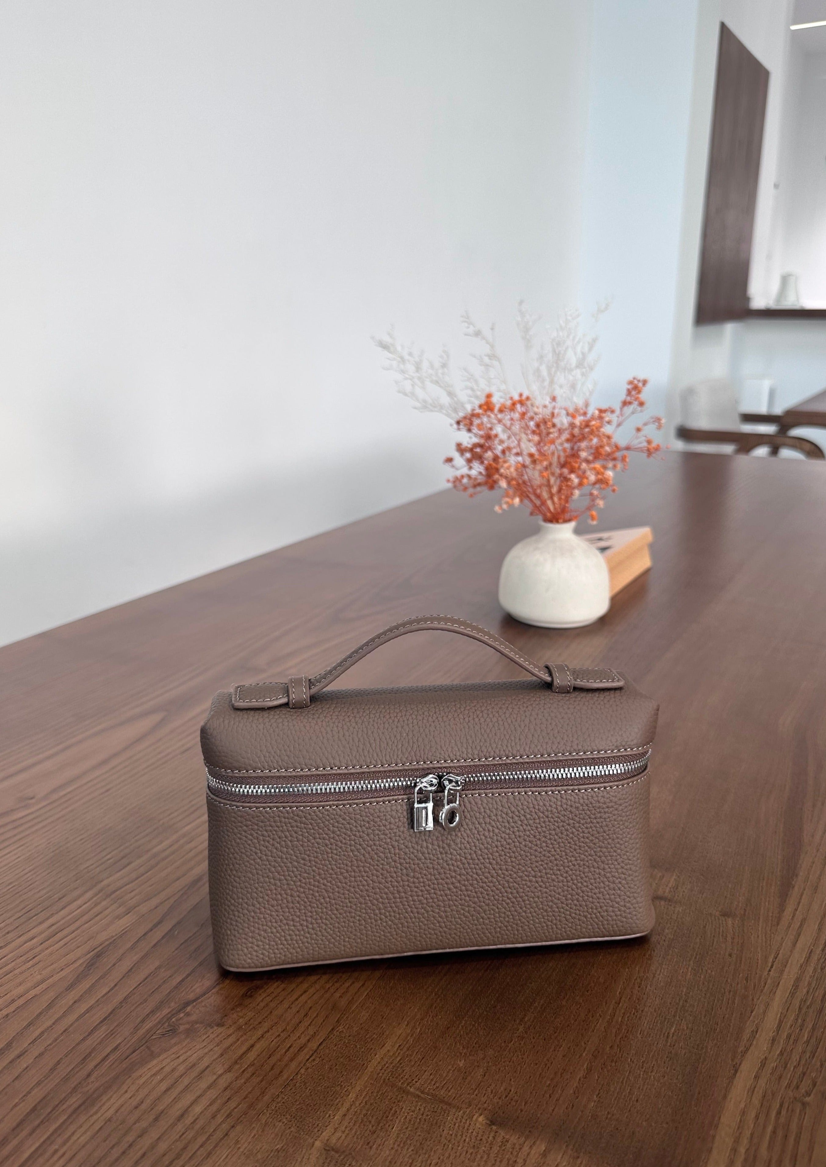 A khaki-colored tote handbag with a long strap placed on a wooden surface, with a small white vase containing dried flowers and a wooden object in the background.