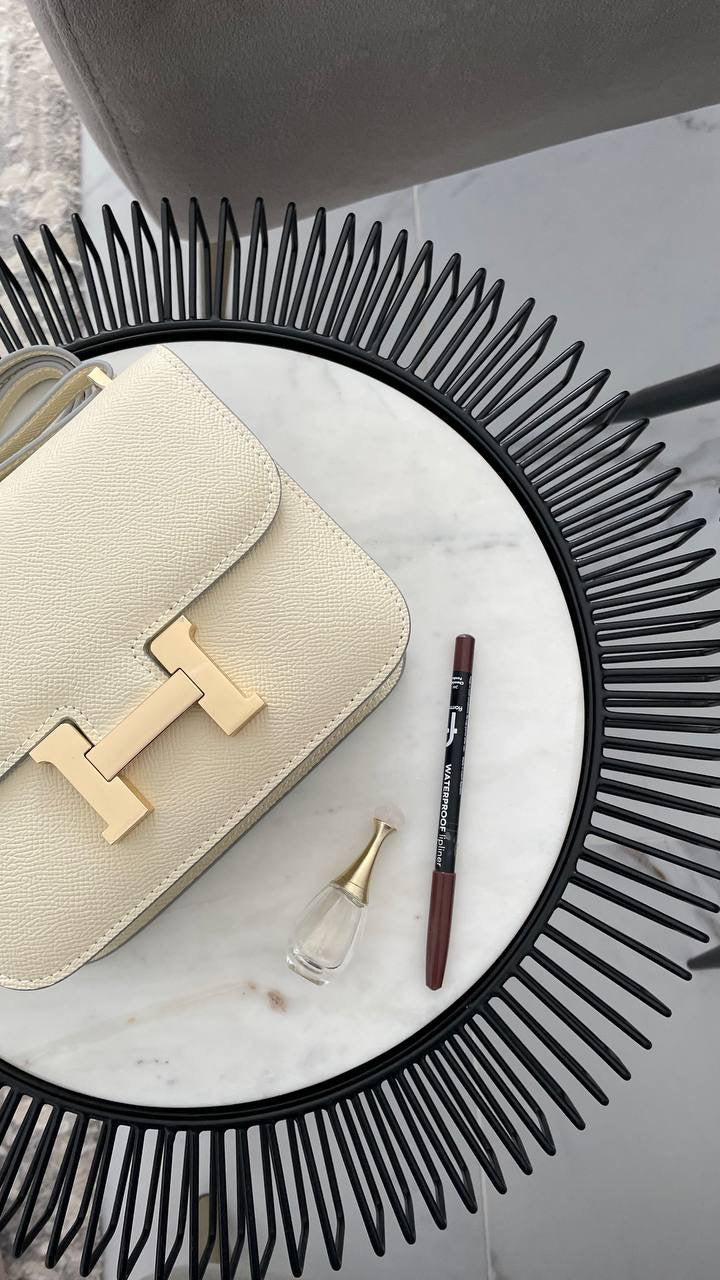 Beige handbag with gold hardware, a small perfume bottle, and two lipsticks on a marble surface.