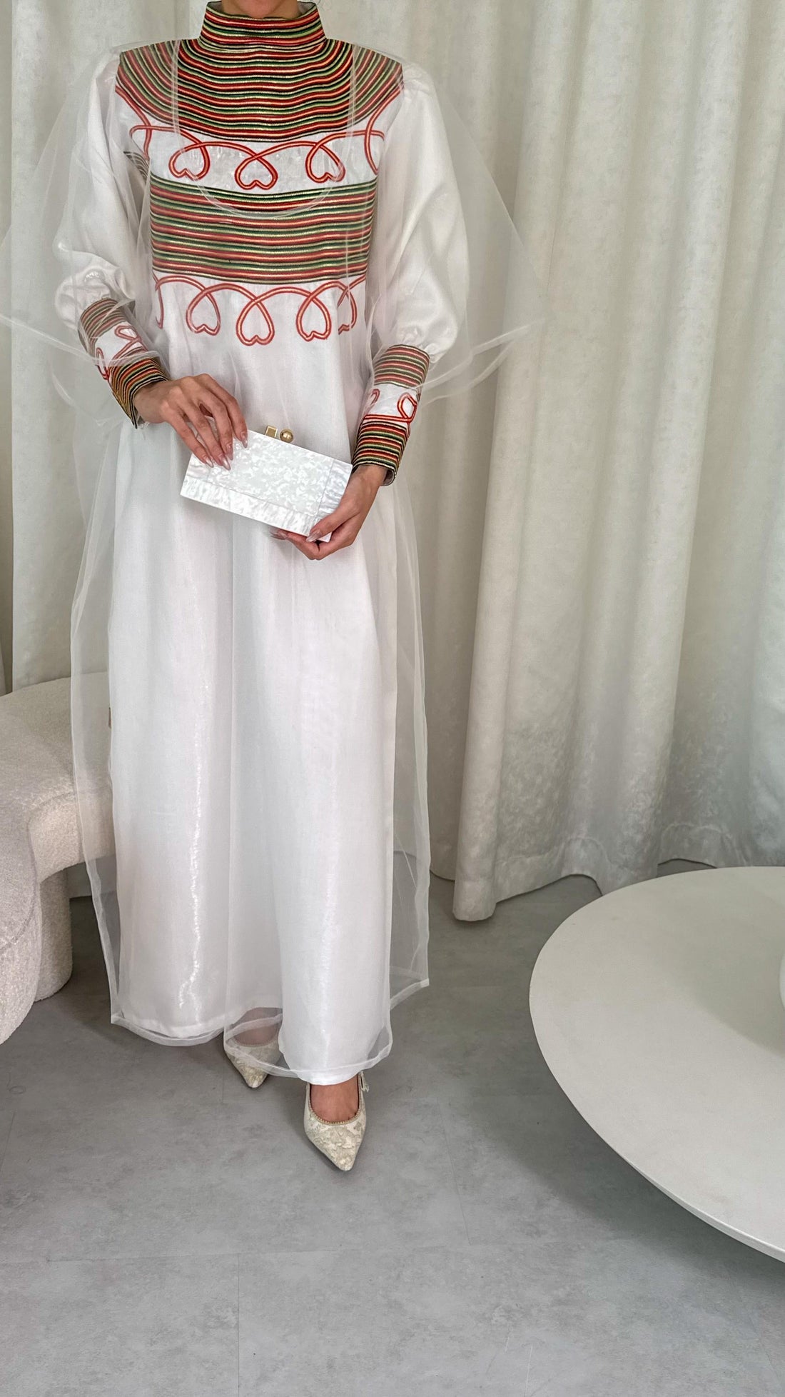 Person wearing a long, light-colored dress with intricate patterns in a minimalistic setting.