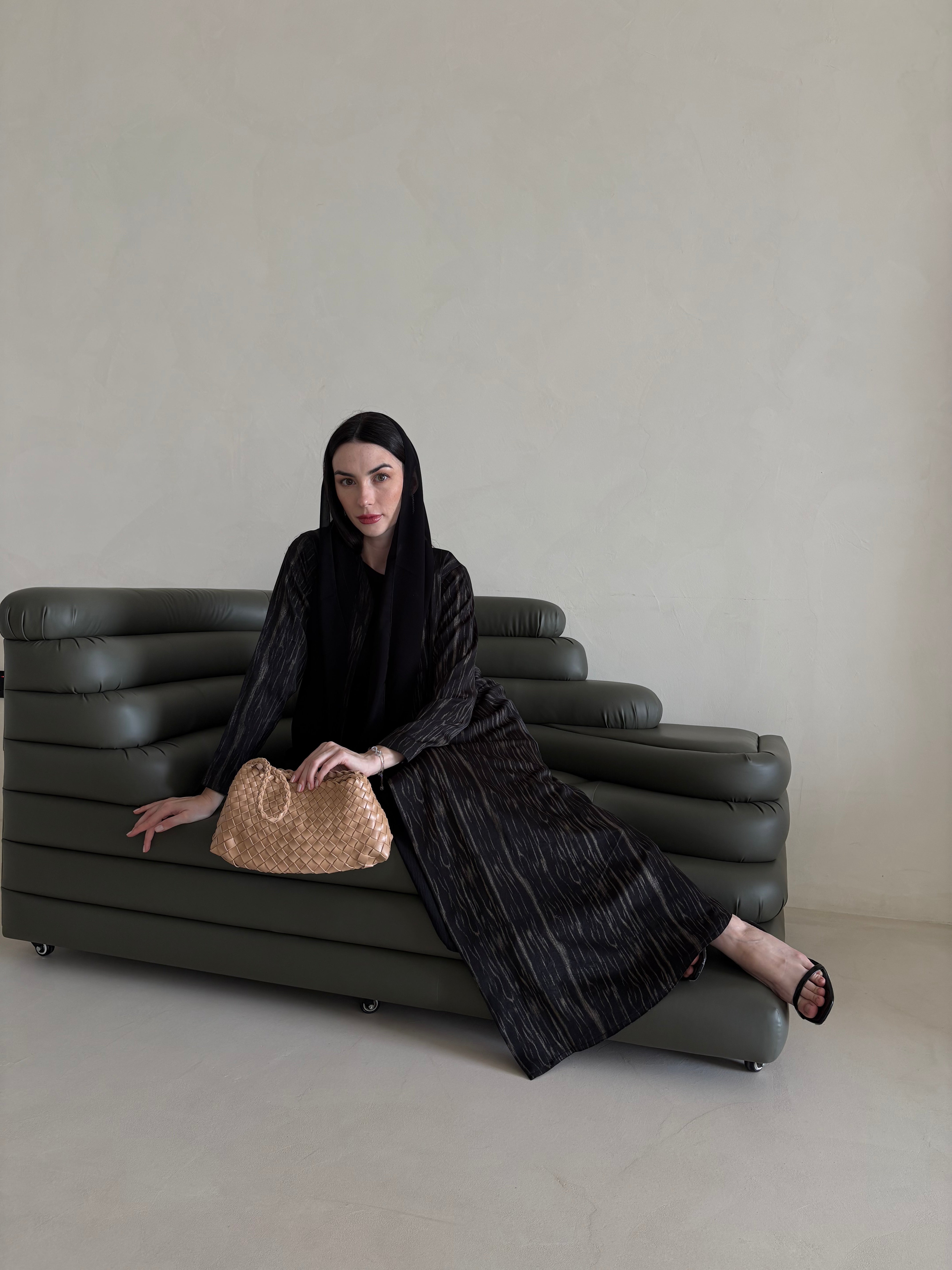 Woman in black outfit sitting on a gray sofa holding a woven bag.