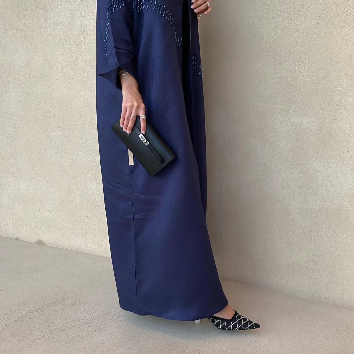 Person wearing a long navy blue dress holding a black clutch against a neutral background