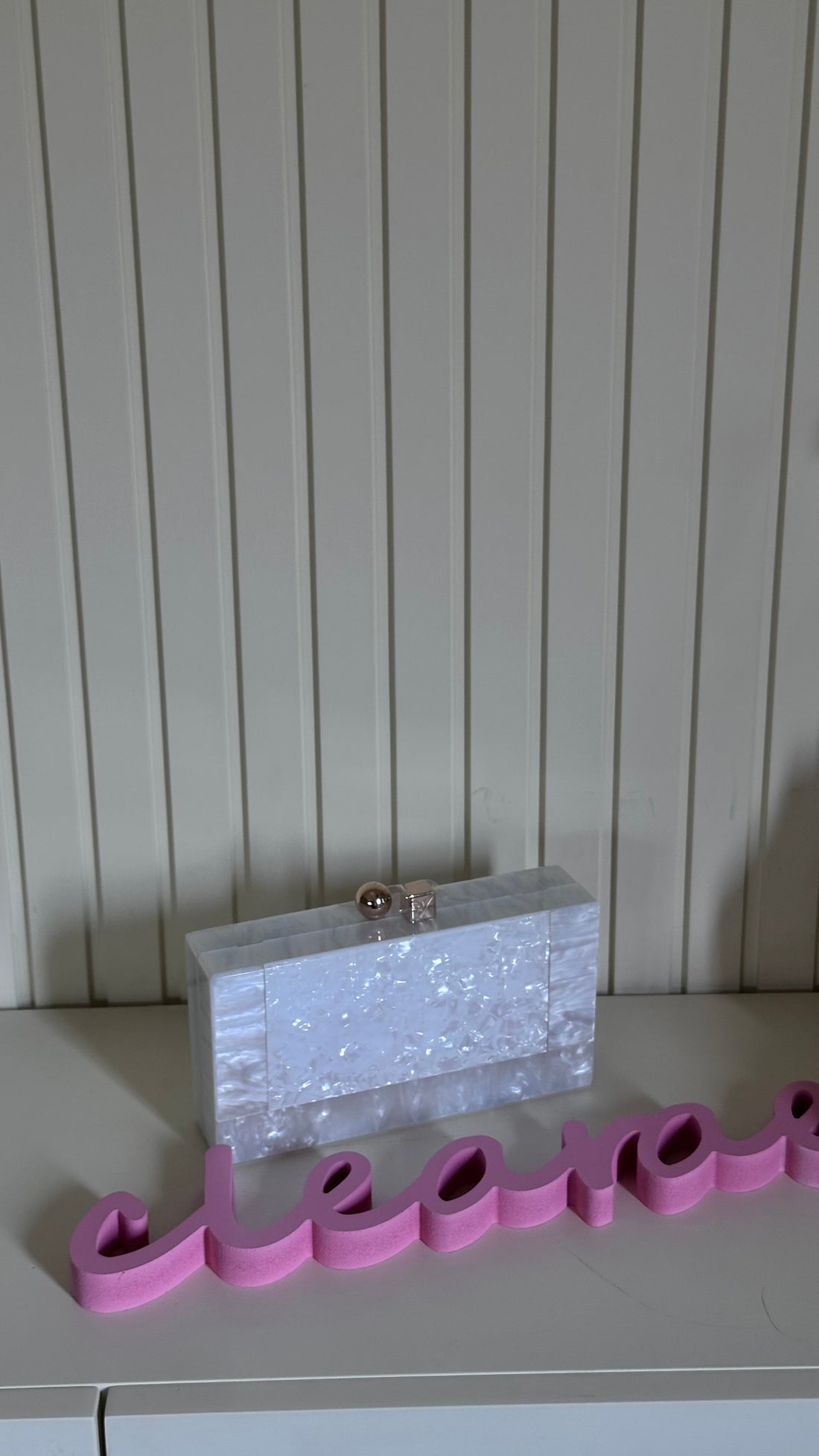 Silver clutch bag on a surface with pink letters spelling 'hello' against a white paneled wall.