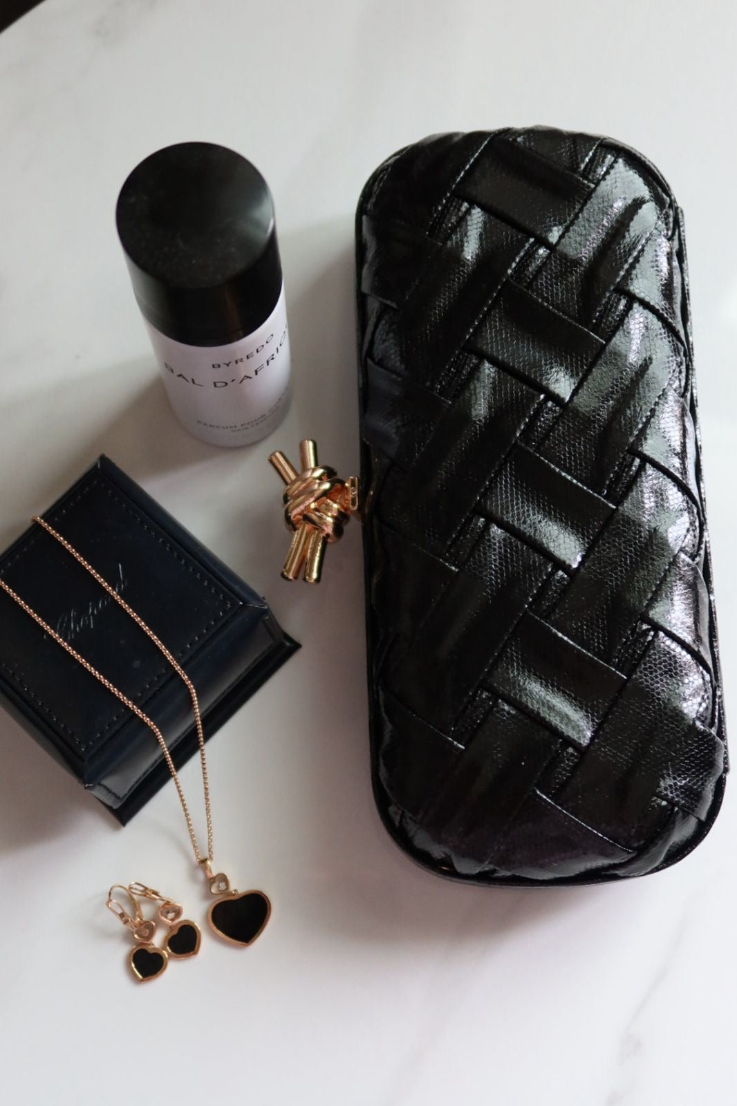 Black woven pouch, white bottle, black jewelry box, gold necklace, and earrings on a light surface.